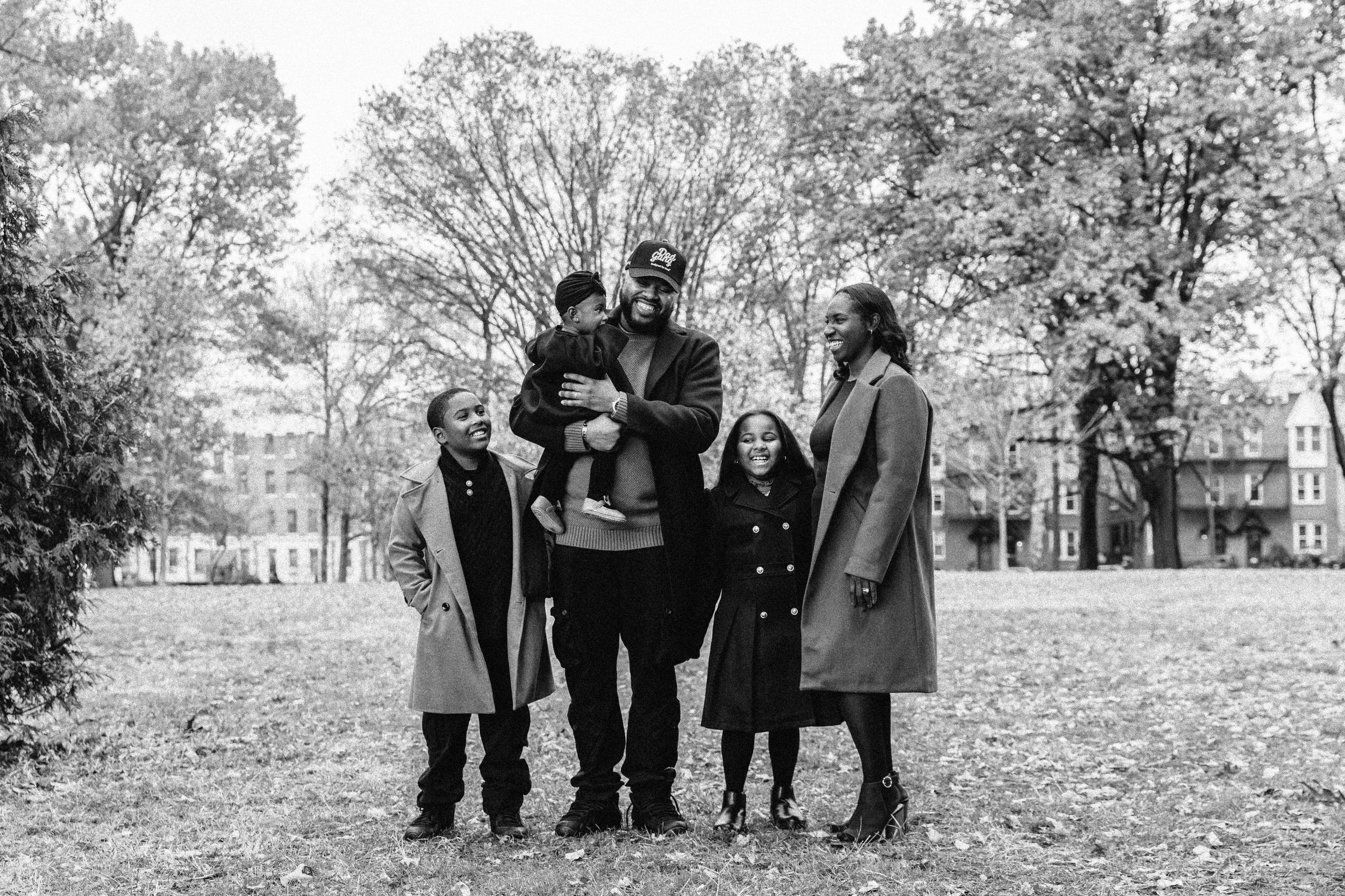 A smiling African American family of five standing outdoors in a park with trees and buildings in the background. Lifestyle family photography in New York City
