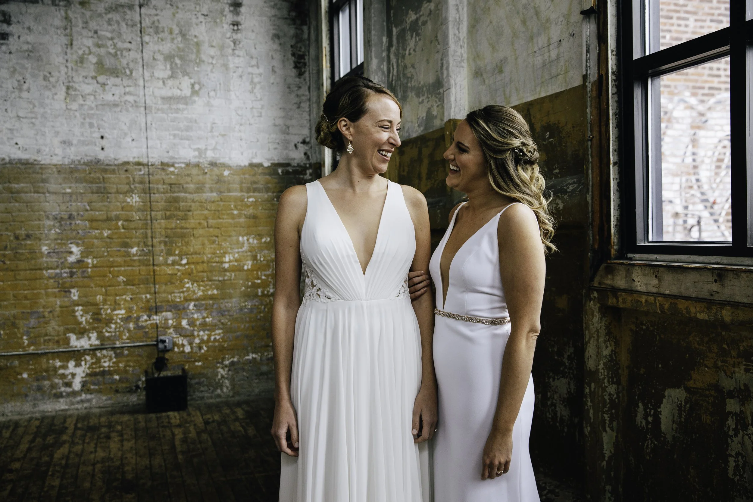 Two women in white dresses smiling and looking at each other in front of a rustic brick wall and window.