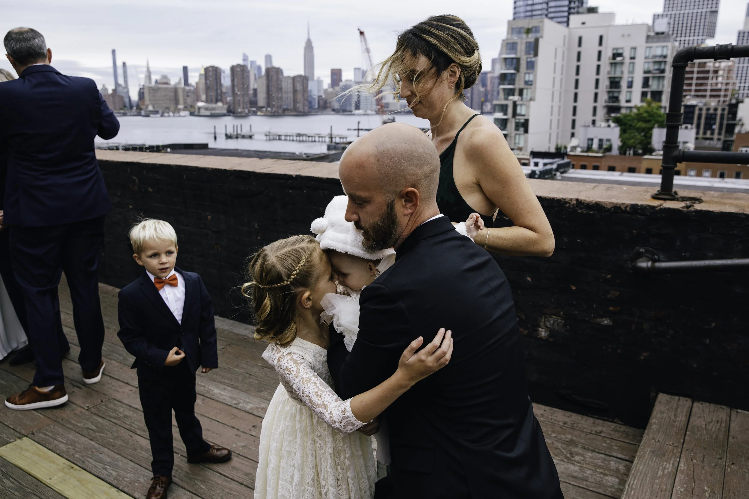 People at a rooftop gathering, including two children in formal attire, with a city skyline in the background. Documentary wedding photography in NYC