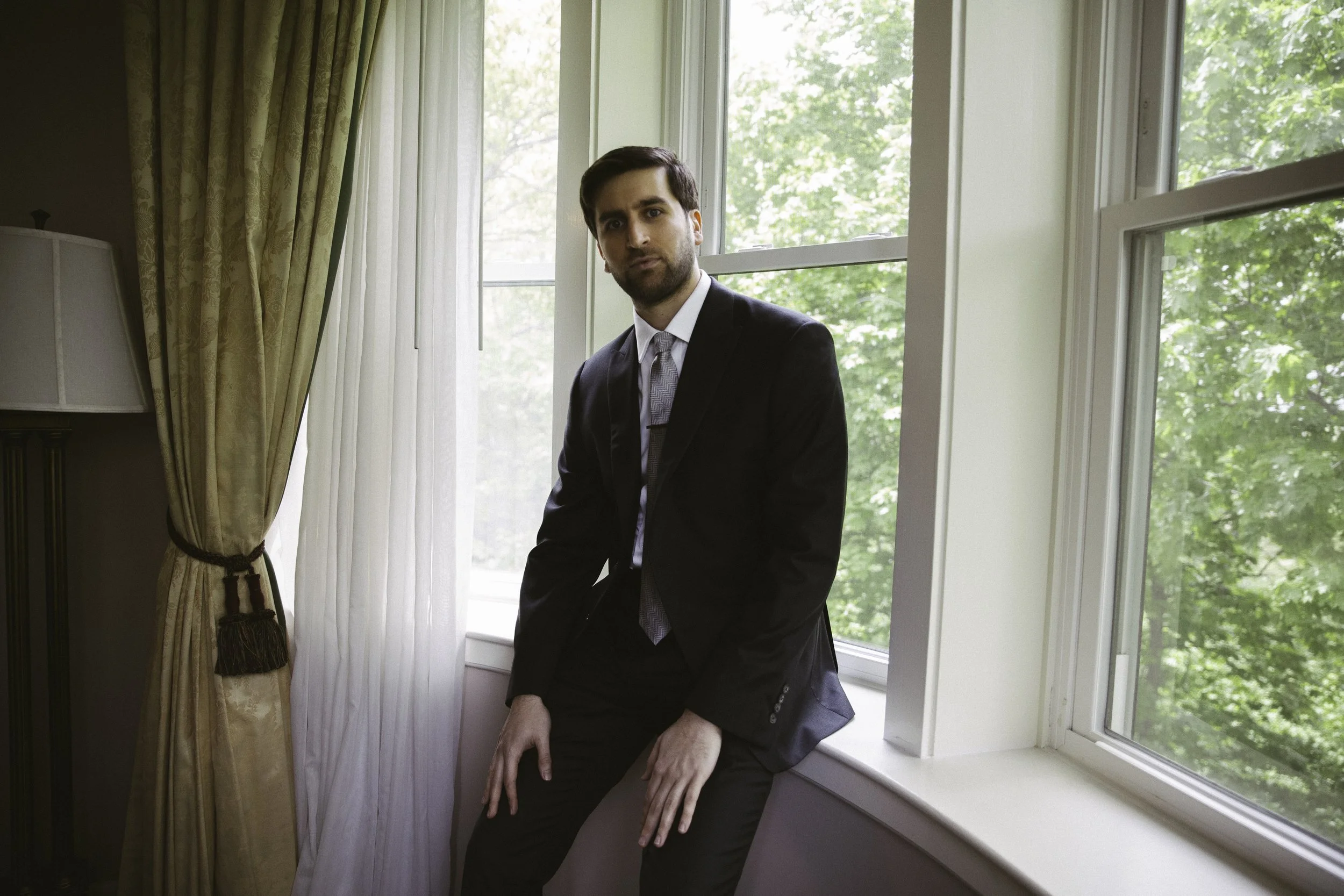A man in a black suit and tie sitting on a window sill in a room with large windows and green trees outside.
