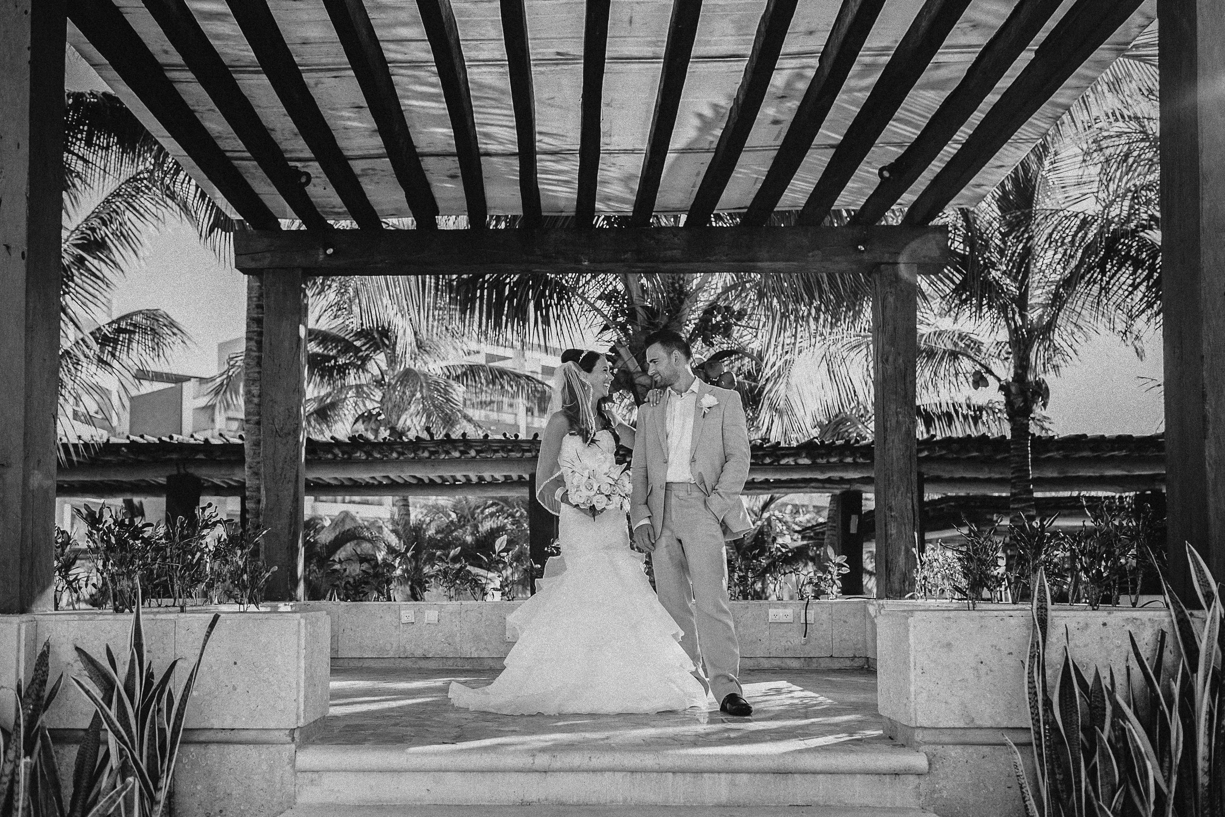 A black and white photo of a bride and groom standing together on a raised platform, surrounded by tropical plants and palm trees, with a wooden pergola overhead. The bride is holding a bouquet and wearing a wedding dress, while the groom is in a lig