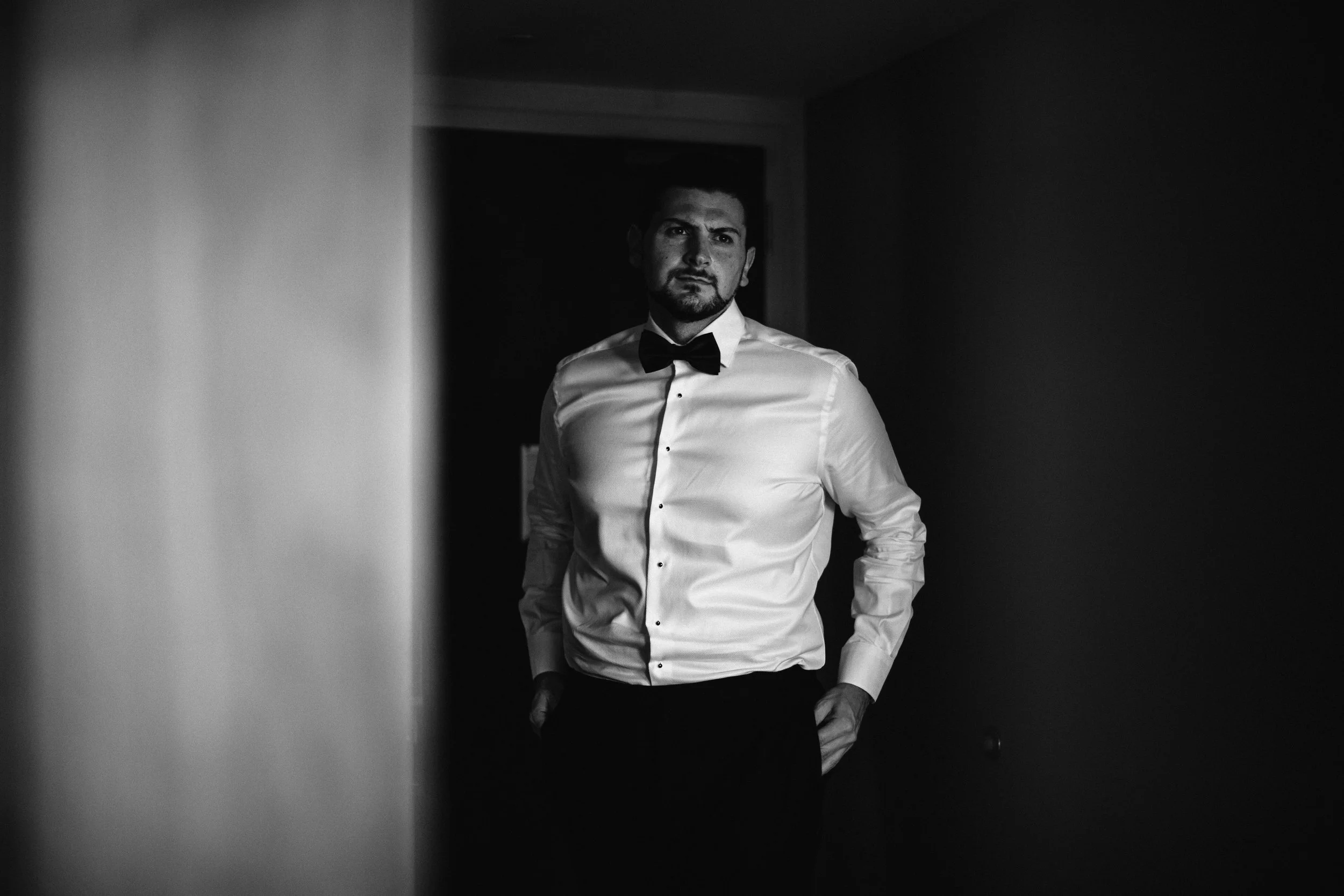 A man in a tuxedo with a bow tie standing in a doorway, looking pensively to the side in a black and white photo. Documentary wedding photography in NYC