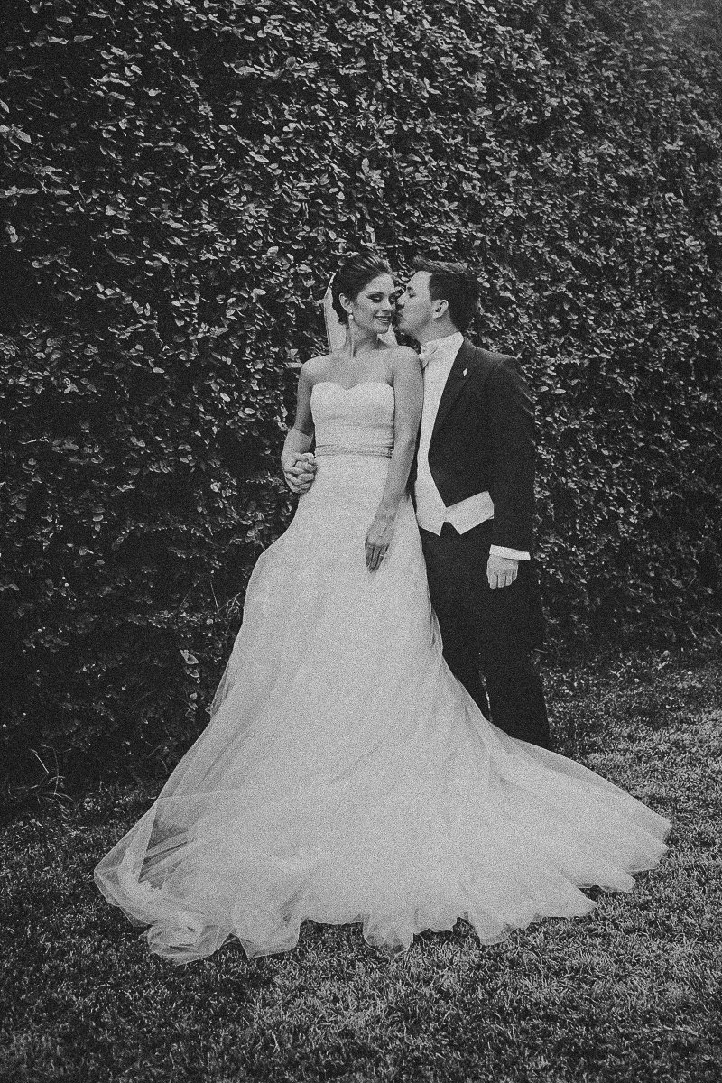 Black-and-white photo of a bride and groom standing outdoors against a leafy hedge, with the groom kissing the bride on the cheek.