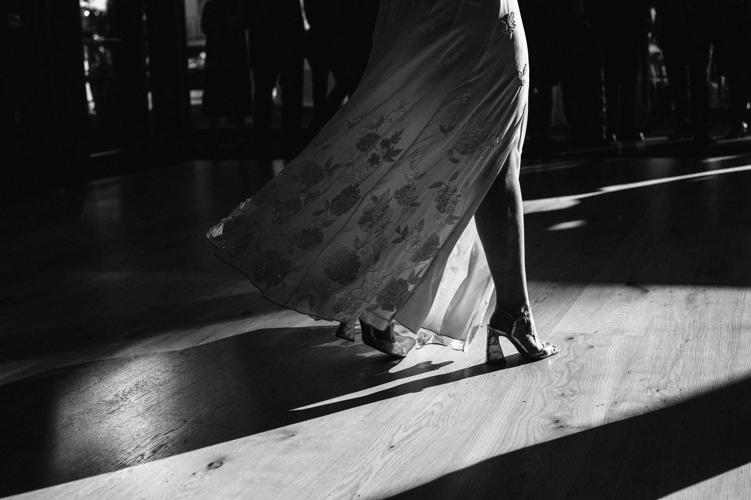 A woman's legs and feet in high heels, walking on a dance floor, with a patterned dress and shadows on the ground. Documentary wedding photography in NYC