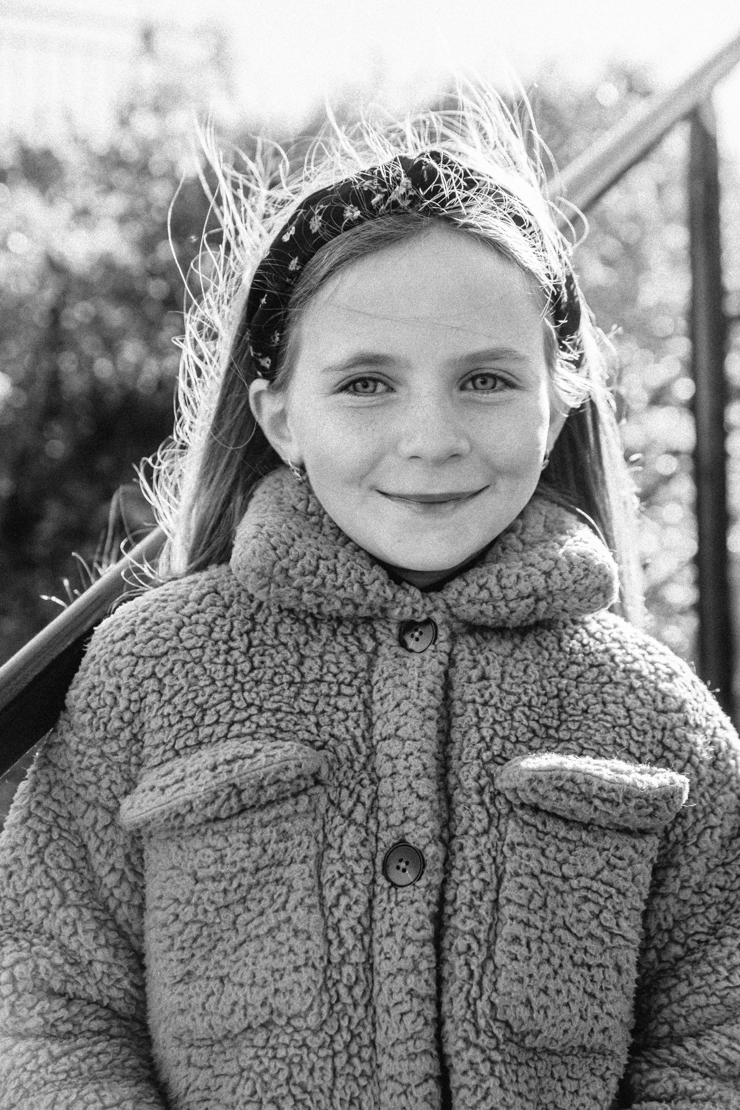 A young girl with long hair, wearing a patterned headband and a fluffy, textured coat, smiling outdoors in sunlight. Documentary portrait photographer in New York City 