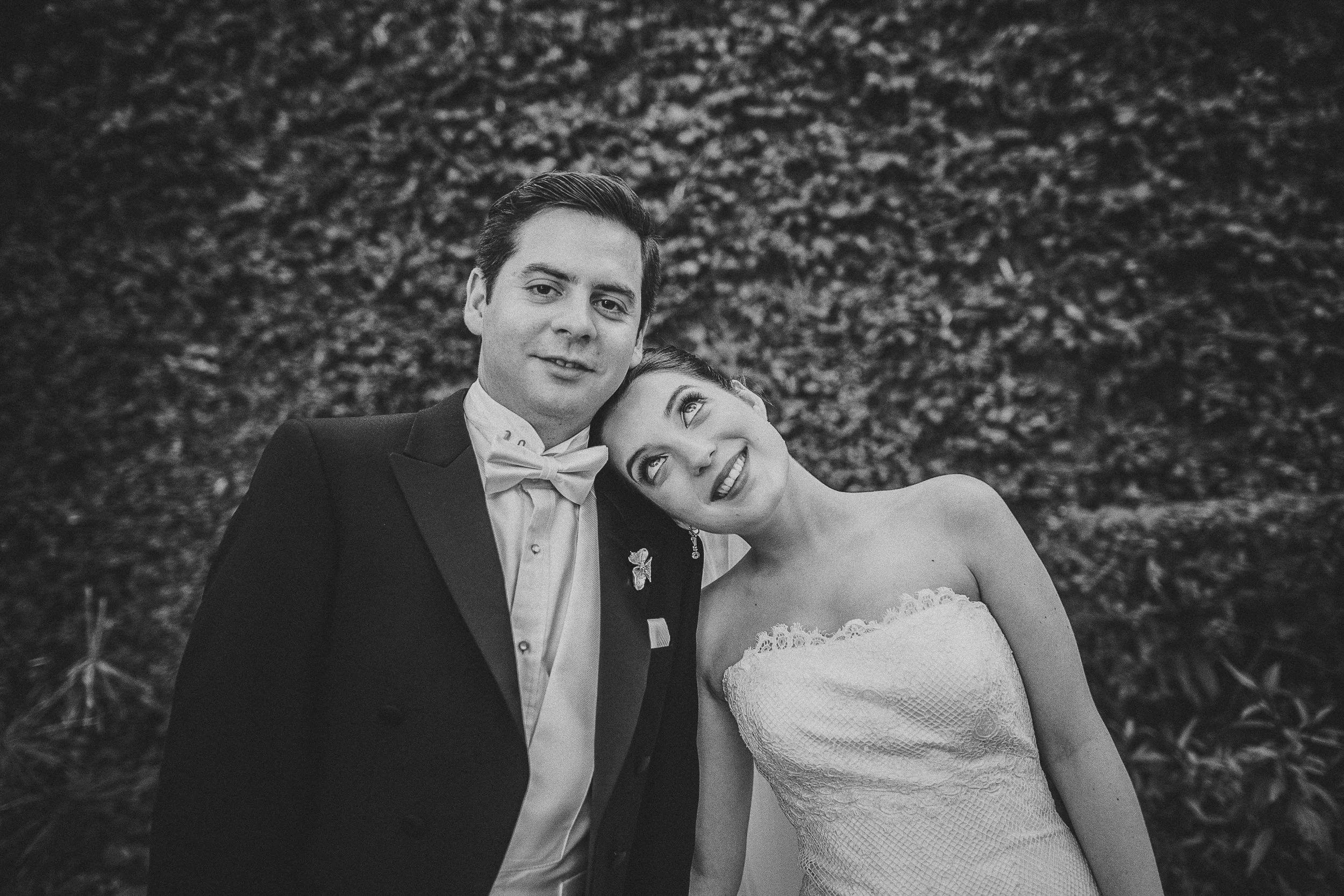 Black and white photo of a happy couple on their wedding day, standing outdoors in front of a leafy background. The groom is wearing a tuxedo with a bow tie, and the bride is in a strapless wedding dress, leaning her head on the groom's shoulder and 