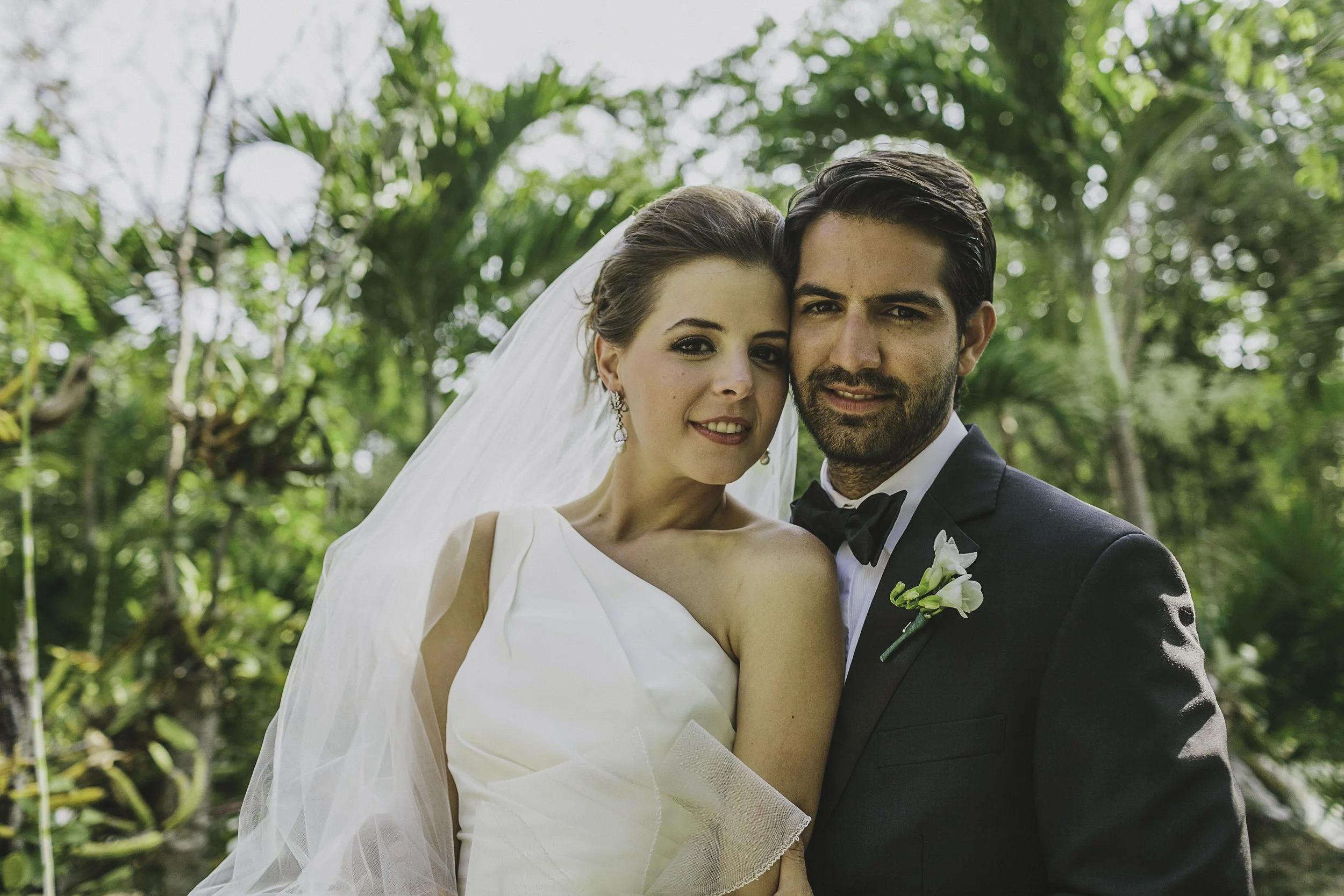 A bride and groom standing close together outdoors, surrounded by green foliage, smiling at the camera.