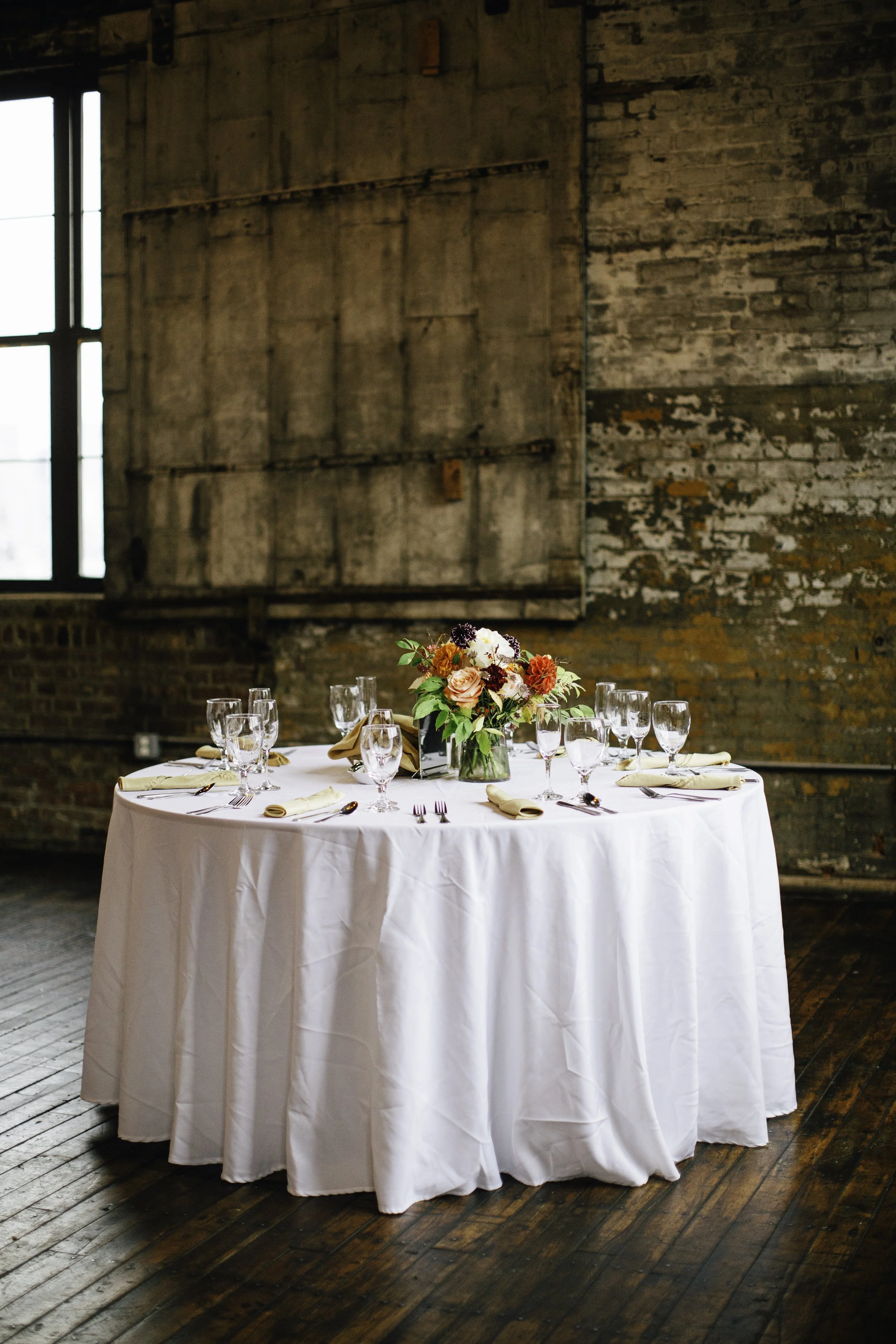 Round table with white tablecloth, set with wine glasses, napkins, silverware, and a bouquet of flowers in the center, in a rustic indoor setting.