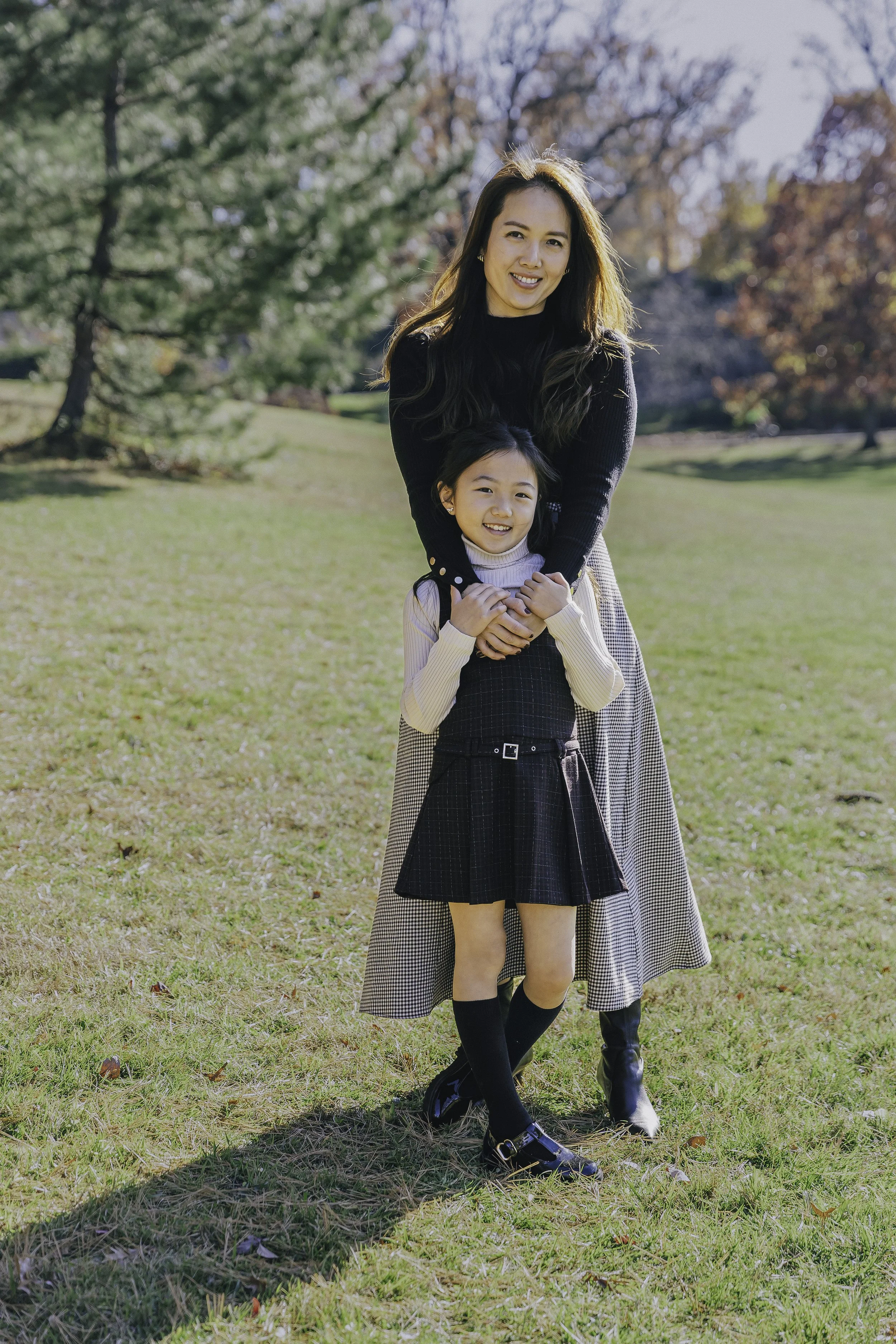A woman and a young girl standing outdoors in a park on a sunny day. The woman has long brown hair and is smiling, wearing a black sweater and a patterned skirt. The girl is smiling, wearing a cream turtleneck, a dark plaid pinafore dress, black sock
