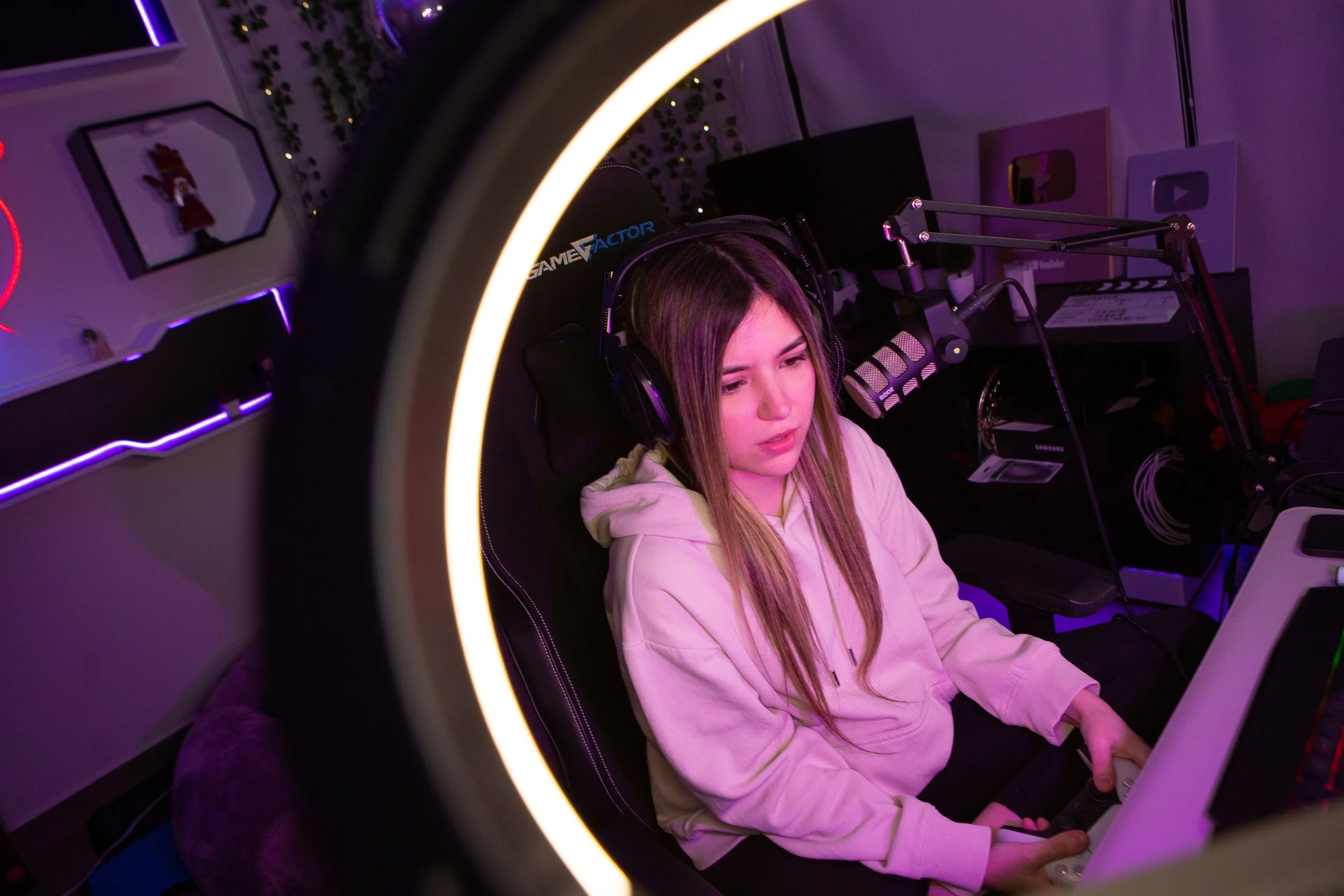 A young woman with long hair wearing a headset and a hoodie sitting in a gaming chair in a dimly lit room, gaming or streaming, with a microphone in front of her and colorful neon lighting.