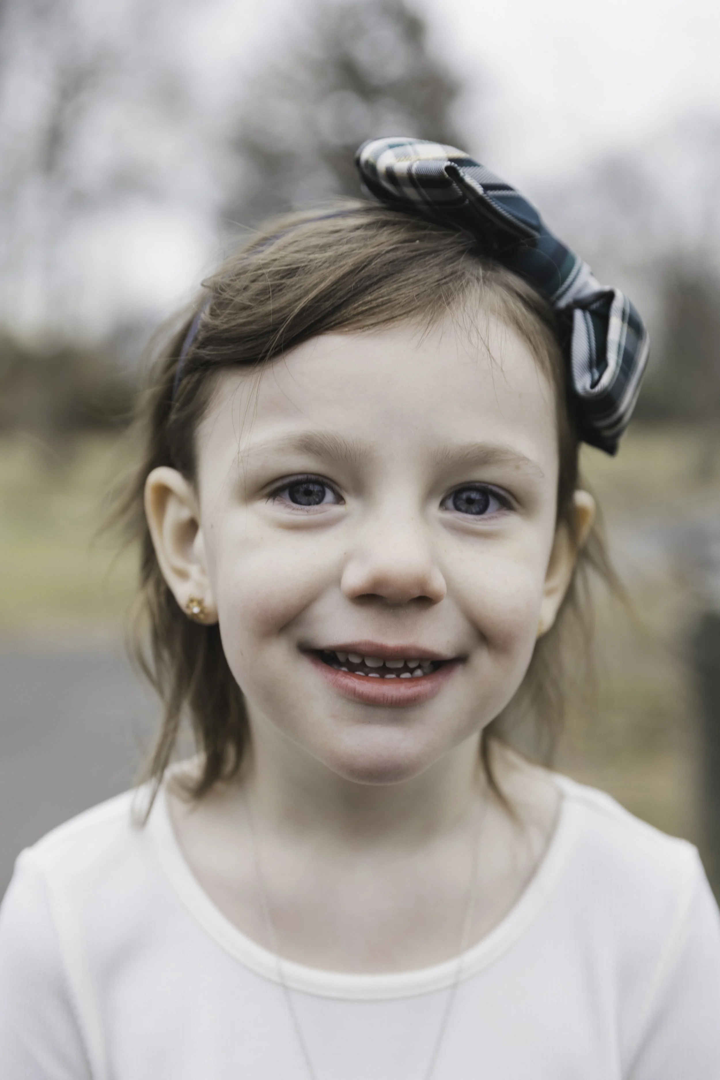 A young girl with blue eyes and brown hair, wearing a white shirt and a large plaid bow in her hair, smiling outdoors.