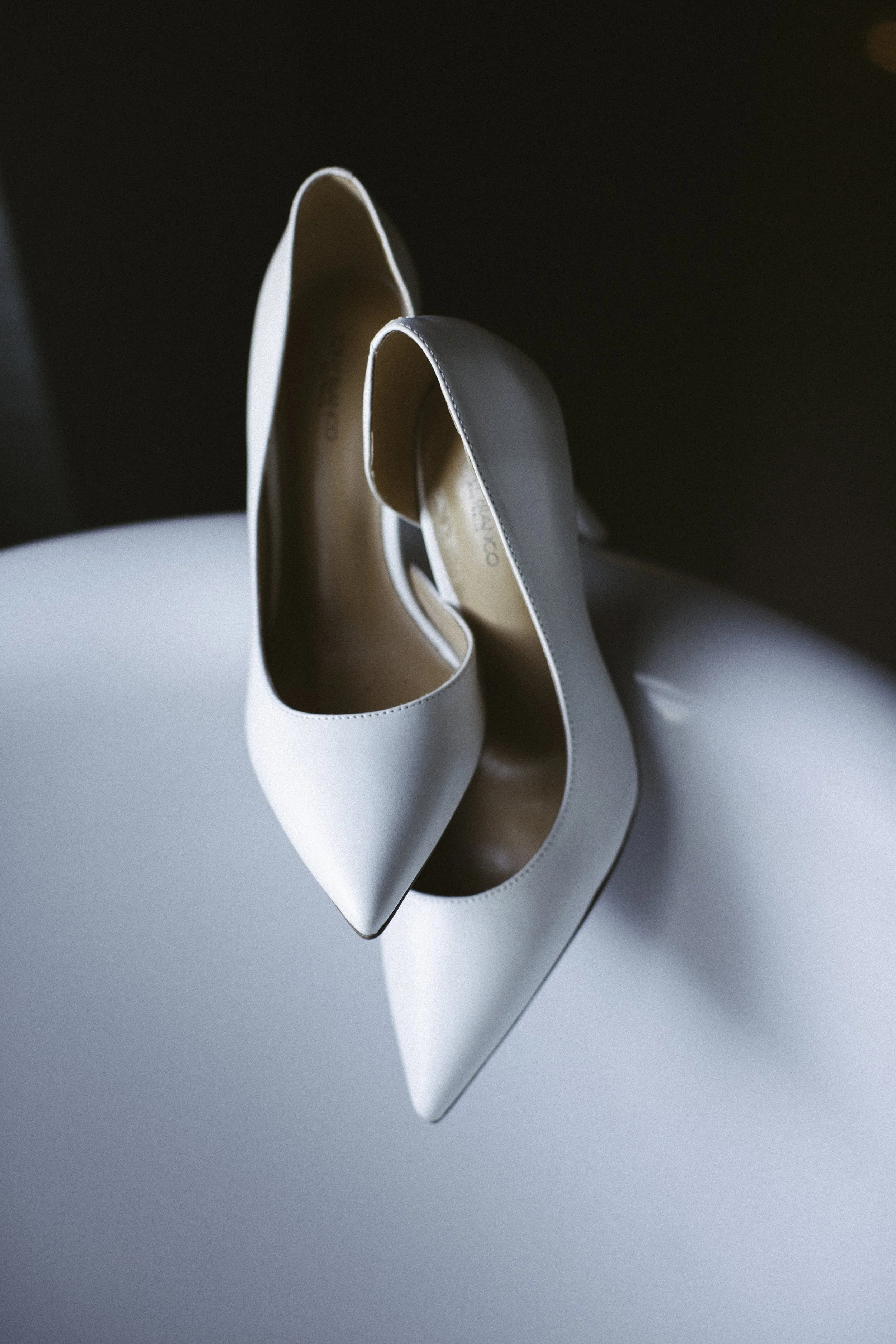 A pair of white high-heeled shoes with pointed toes resting on a white surface. Documentary wedding photography in NYC