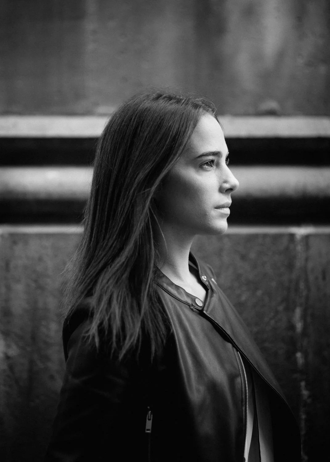 Black and white photo of a young woman with long hair in profile, wearing a leather jacket, with a blurred background.