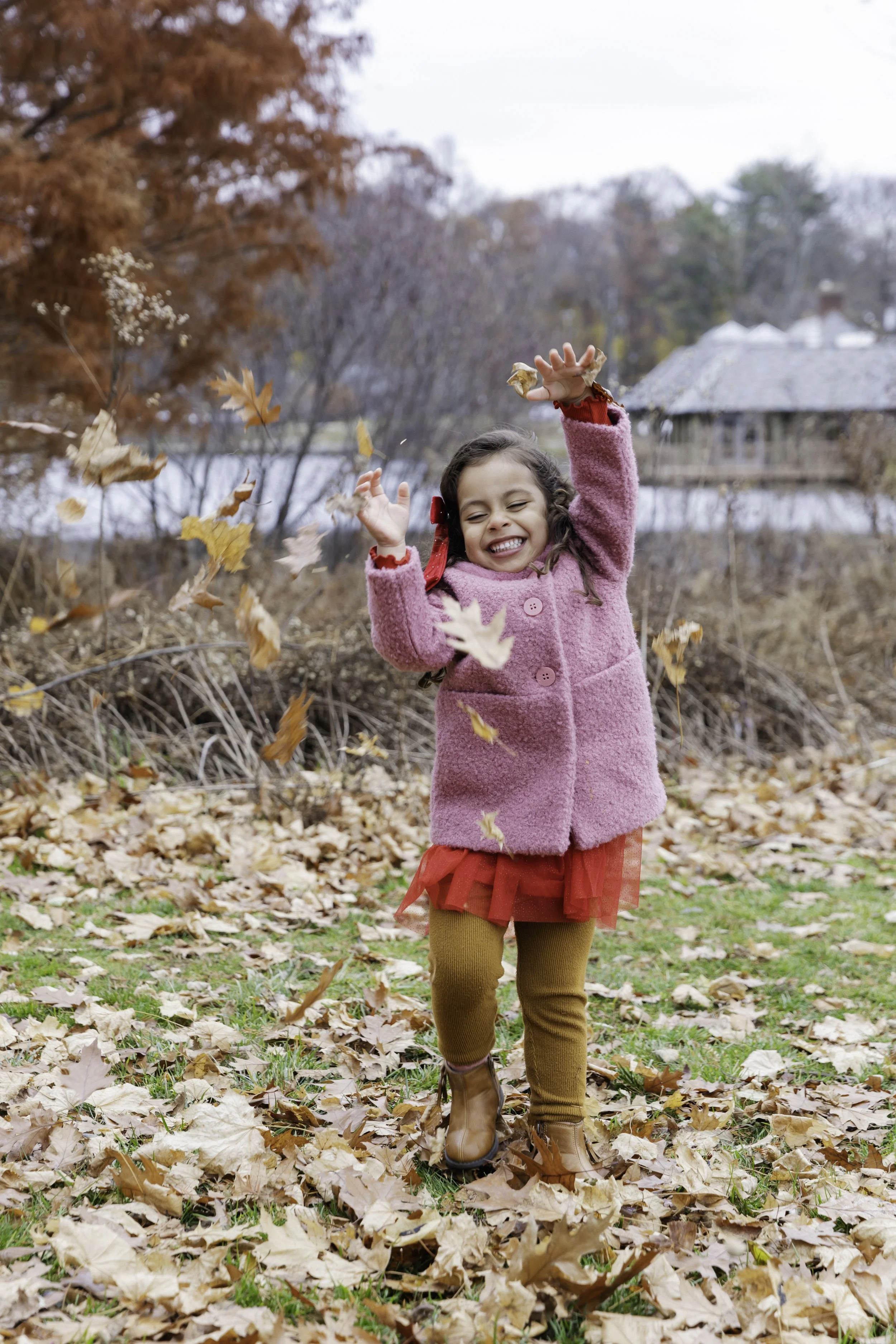 A young girl in a pink coat and red dress playing in fallen autumn leaves in a park, smiling and enjoying fall weather.