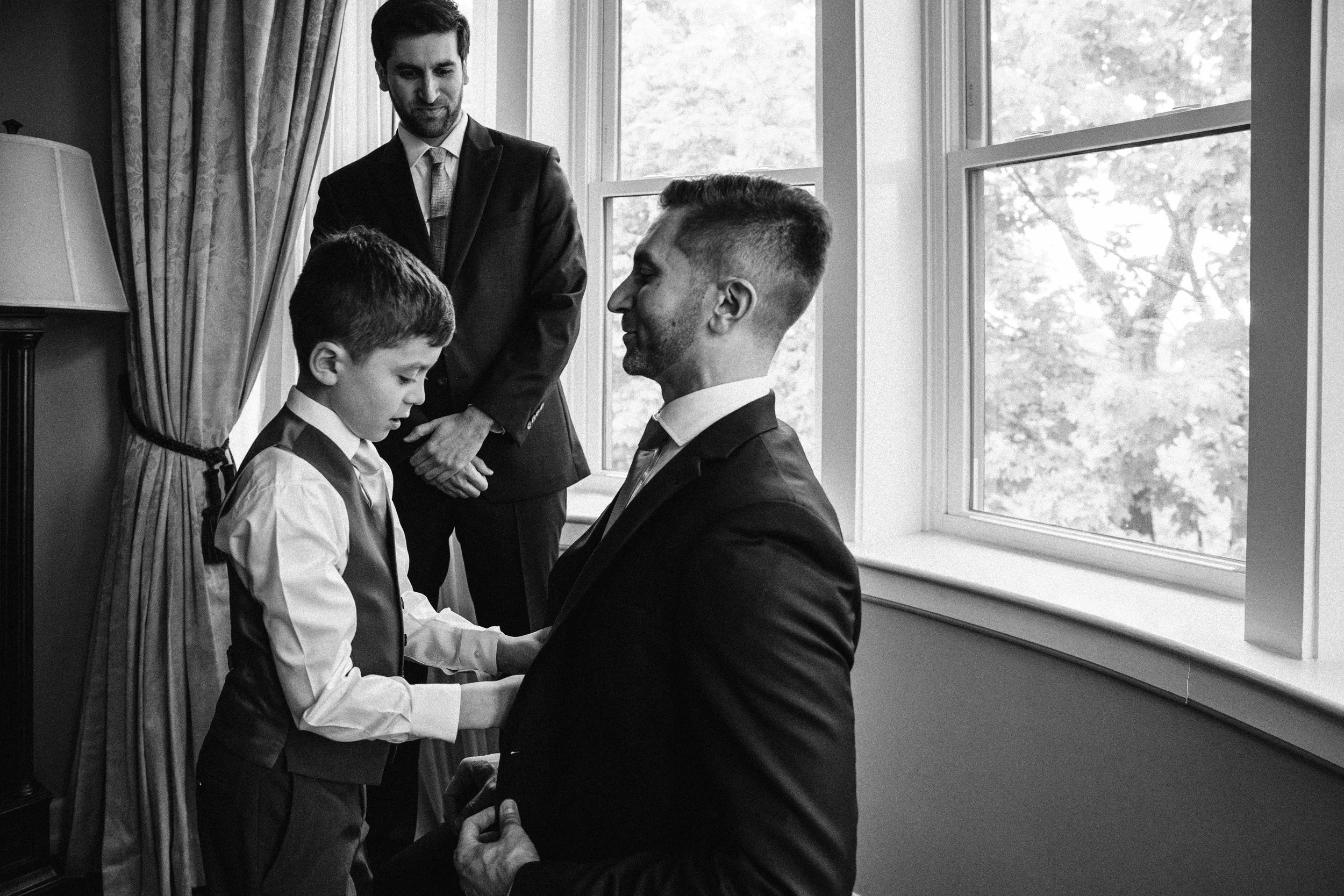 A man in a tuxedo sitting and looking at a young boy, who is also in formal attire, with another man standing behind them in a well-lit room near a window. Documentary wedding photography in NYC