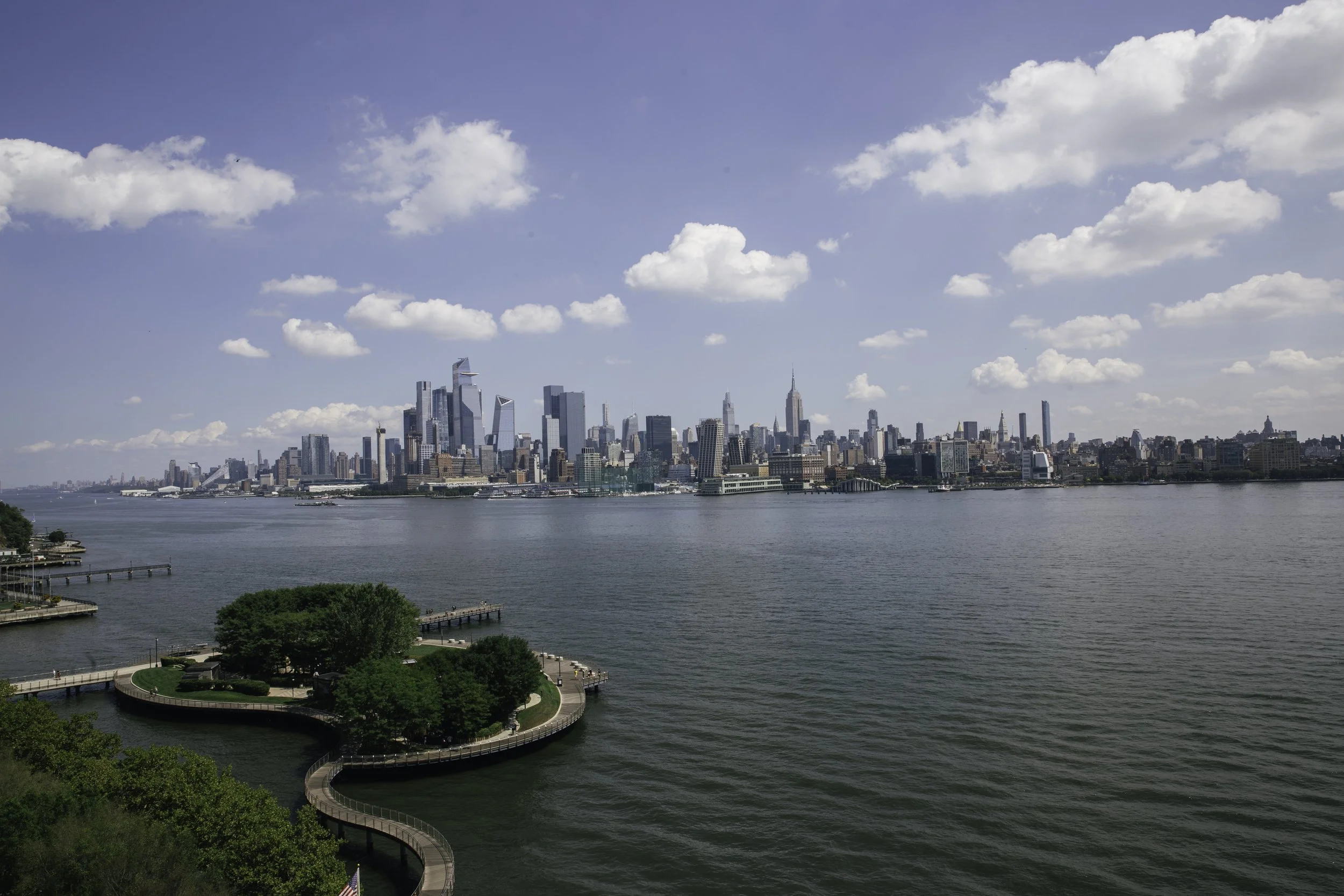 View of the New York City skyline across the Hudson River, featuring skyscrapers and a partly cloudy sky.