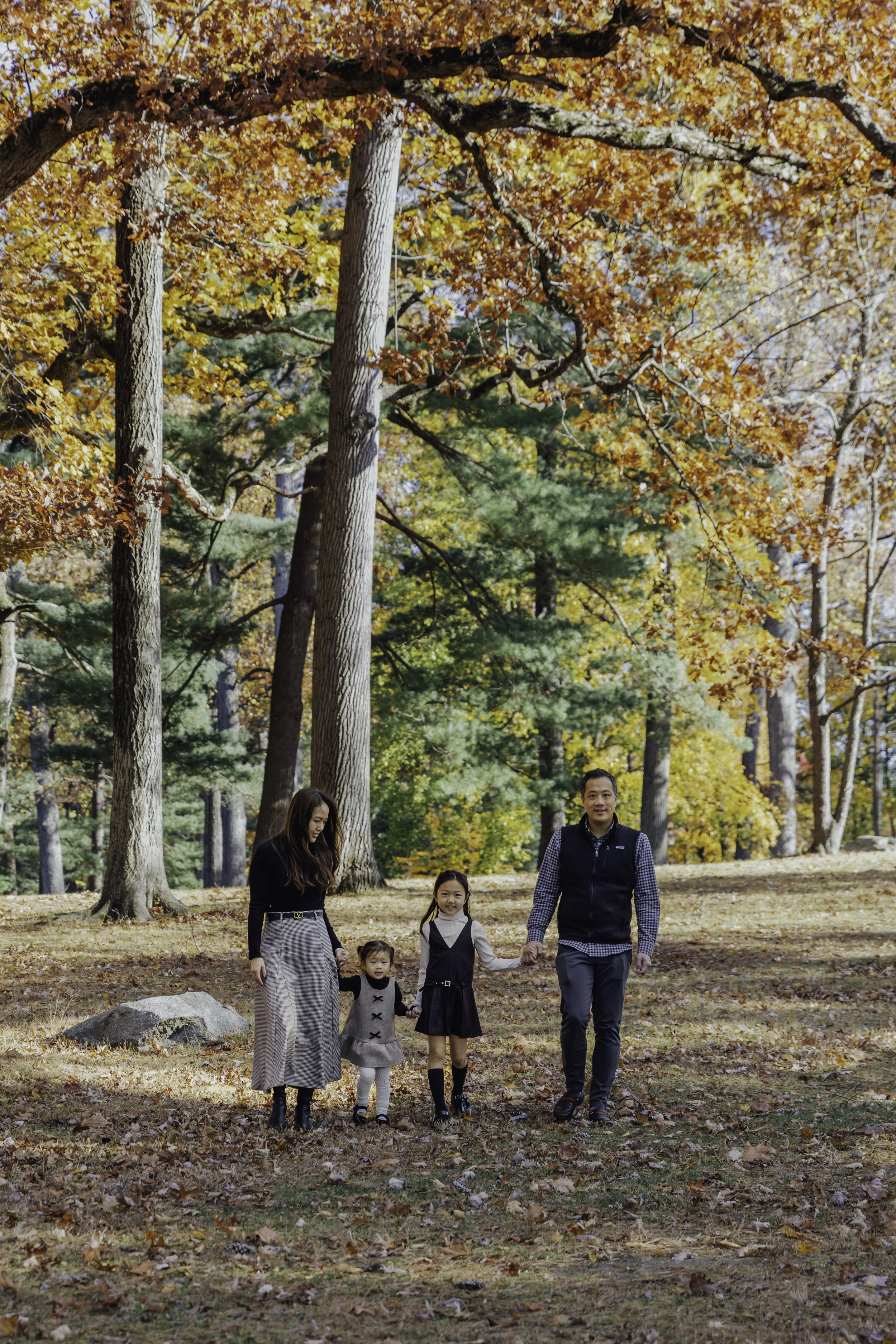 A family of four walking hand-in-hand through a wooded park with colorful autumn leaves. Lifestyle family photography in New York City