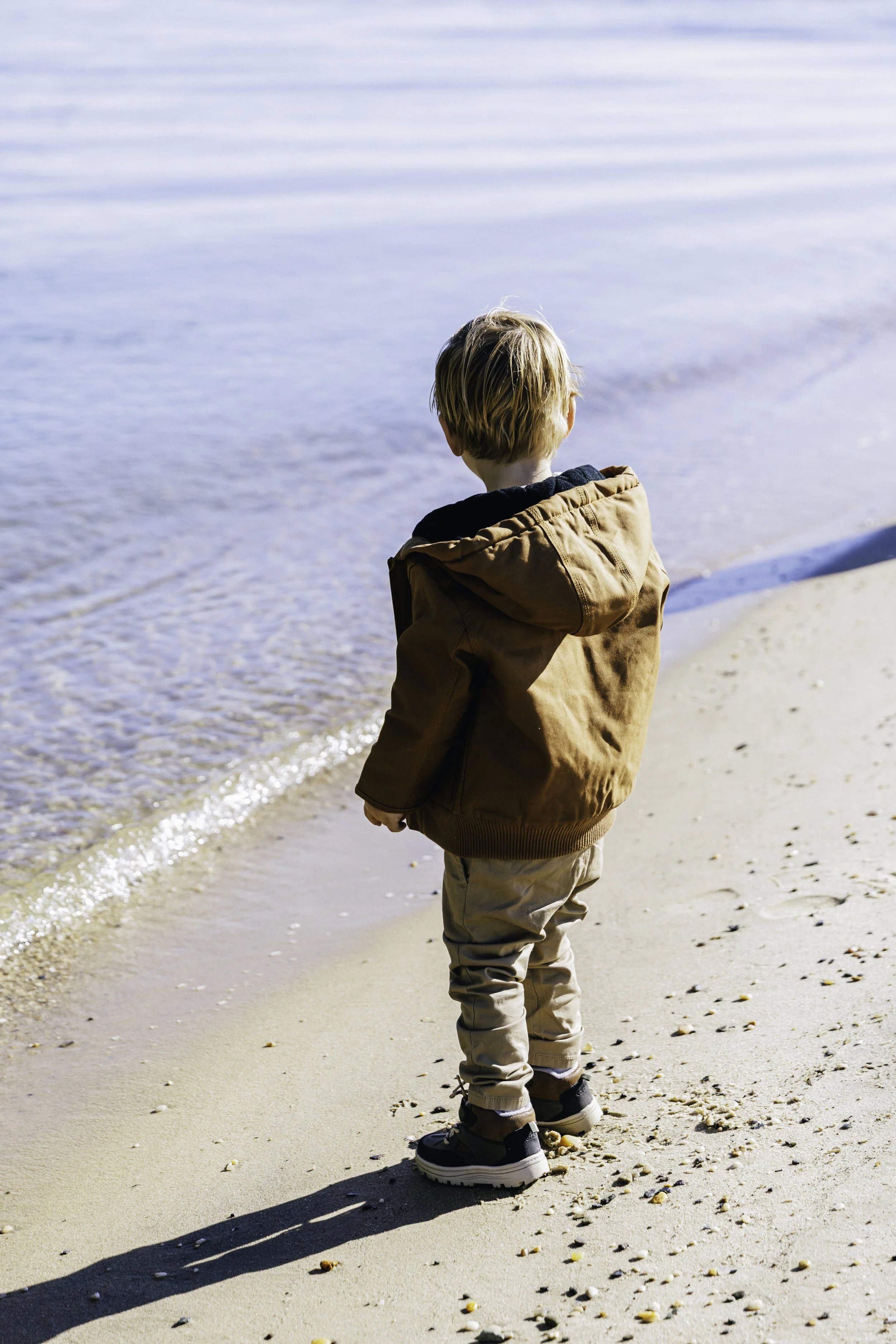 A young boy standing on the sandy beach near the ocean, facing the water, wearing a brown jacket, beige pants, and black sneakers.