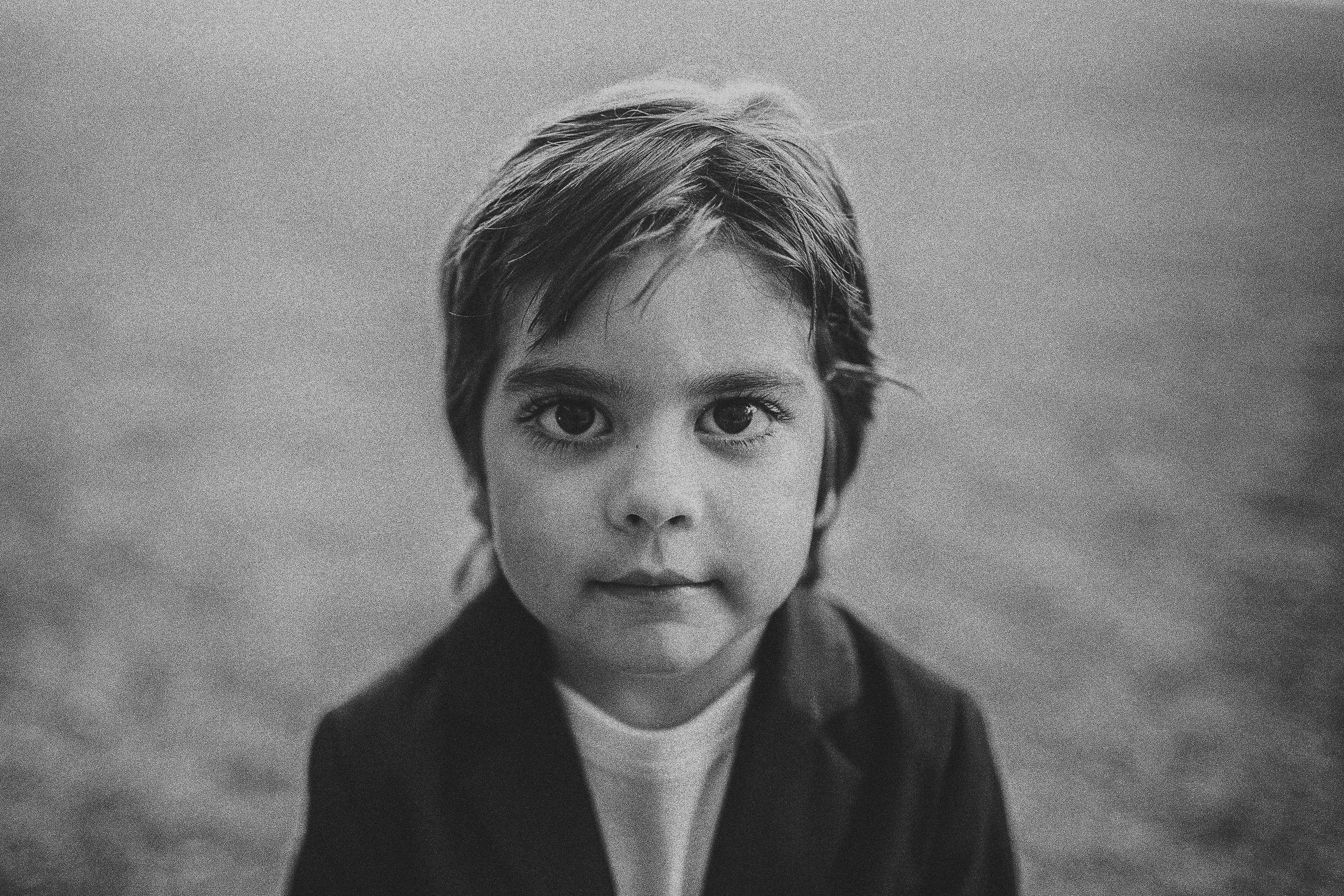 Black and white portrait of a young child with short, tousled hair, wearing a dark jacket and a white shirt underneath, looking directly into the camera.