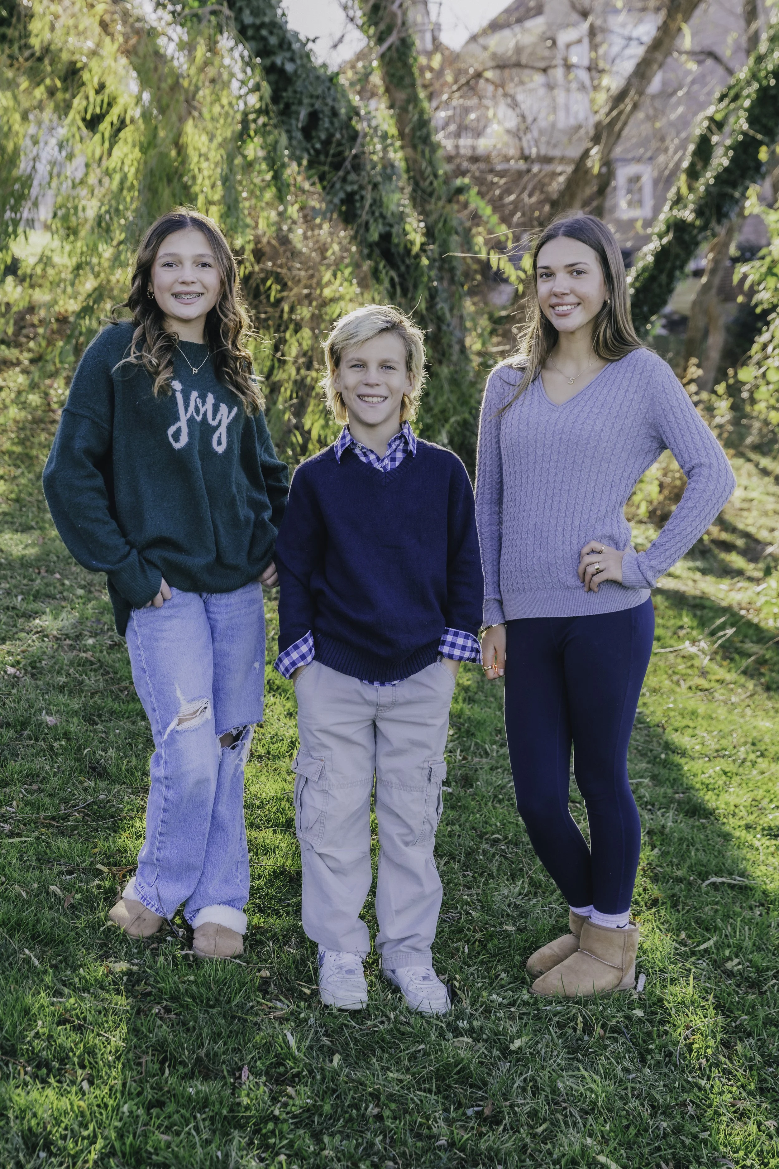 Three kids, two girls and one boy, standing outdoors on grass with trees and sunlight behind them, smiling at the camera. Lifestyle family photography in New York City