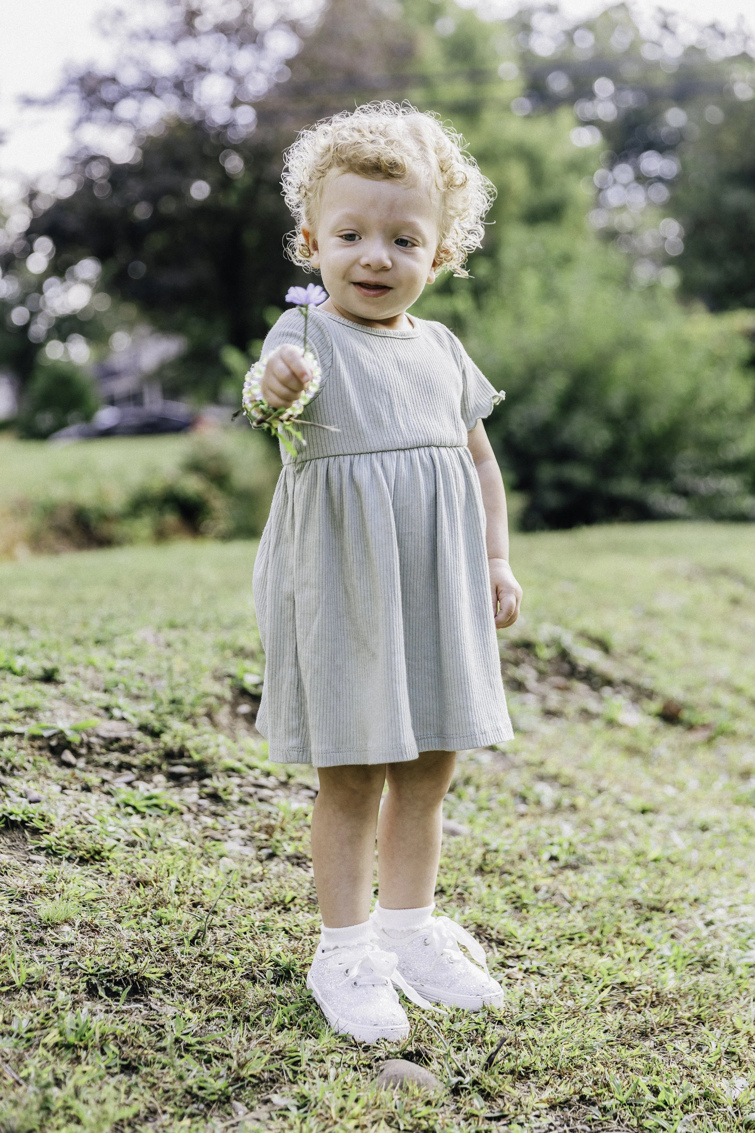 A young girl with curly blonde hair standing on grass in a park, holding a small flower. She is wearing a light gray dress, white socks, and white shoes, with trees and a house in the background.