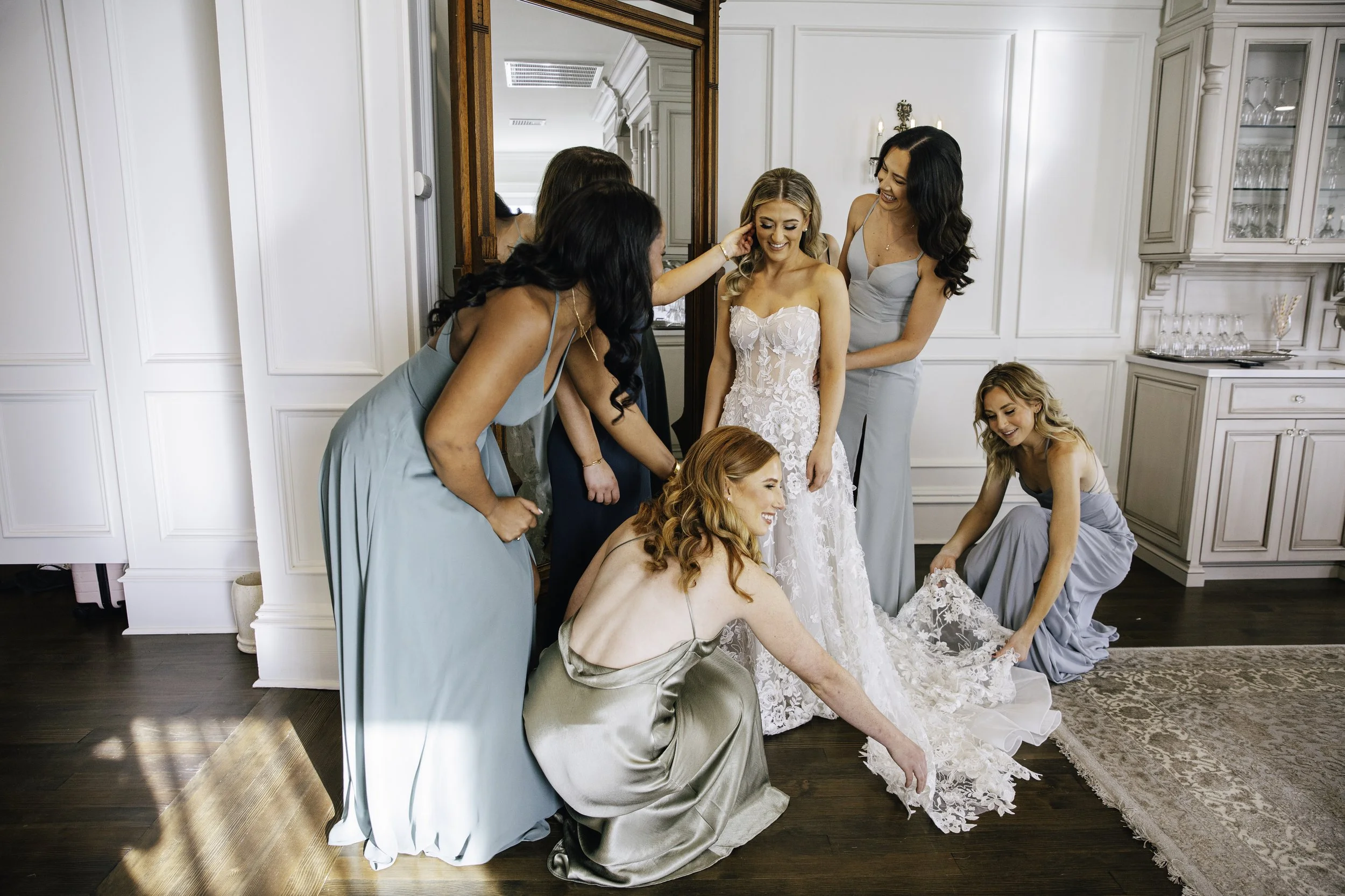A bride with blonde hair in a white lace wedding gown is surrounded by her bridesmaids helping her prepare for the wedding in a bright, elegant room. Documentary wedding photography in NYC