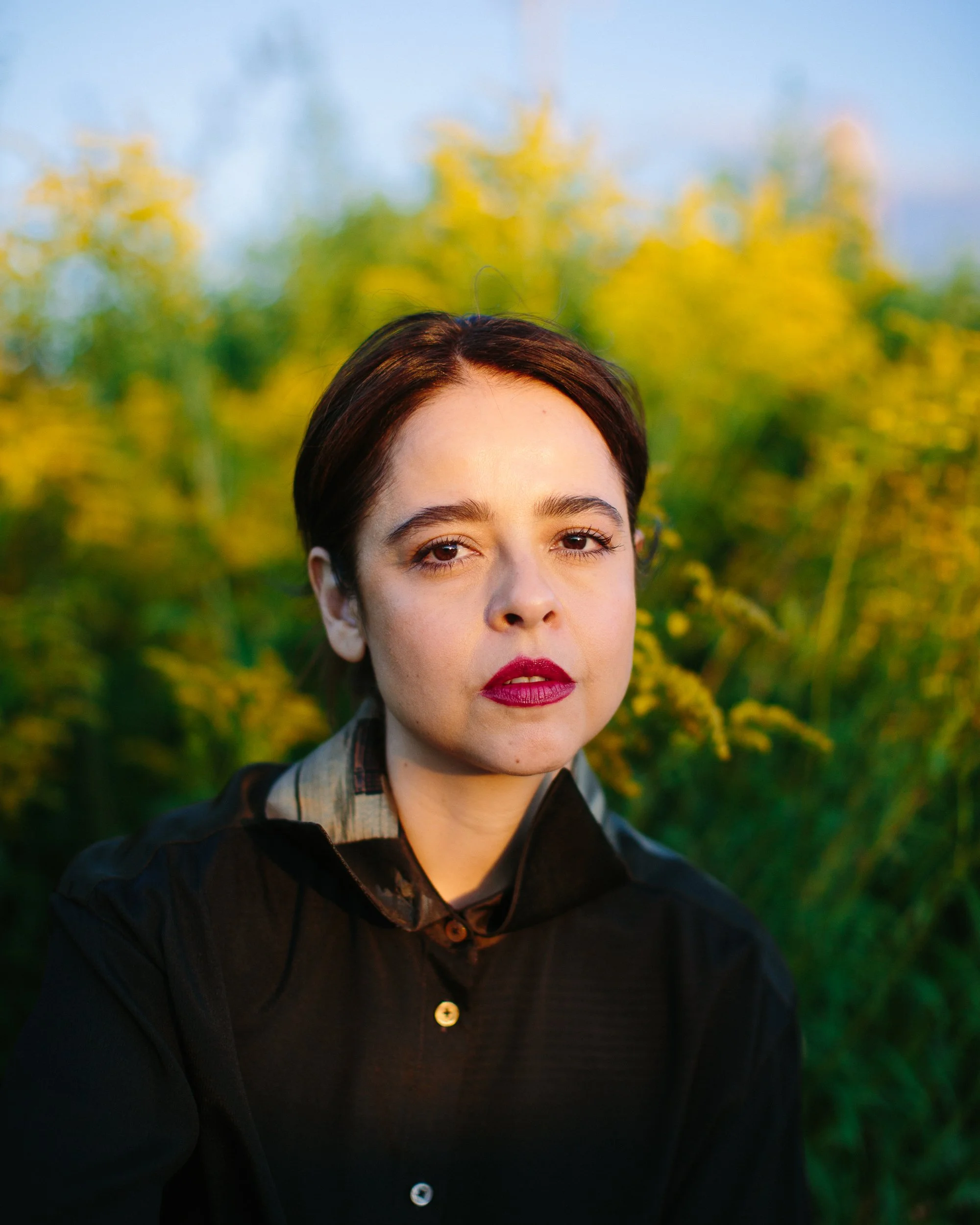 A woman with short dark hair, wearing a black top, standing outdoors among yellow flowering plants, with a background of trees and a blue sky.