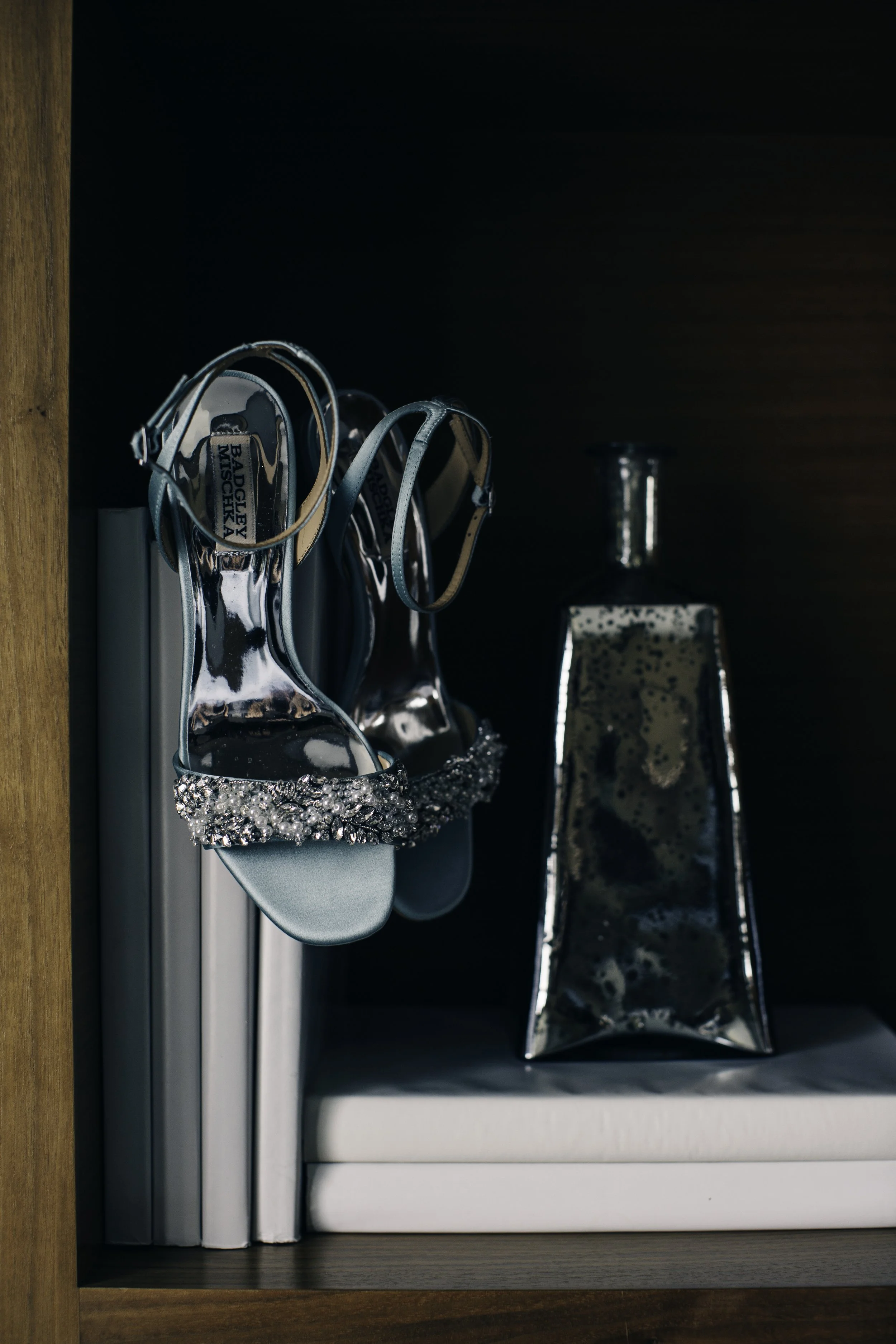 A pair of silver high-heeled sandals with rhinestones on the straps, hanging from a corner of a bookcase, with a metallic decorative vase nearby.