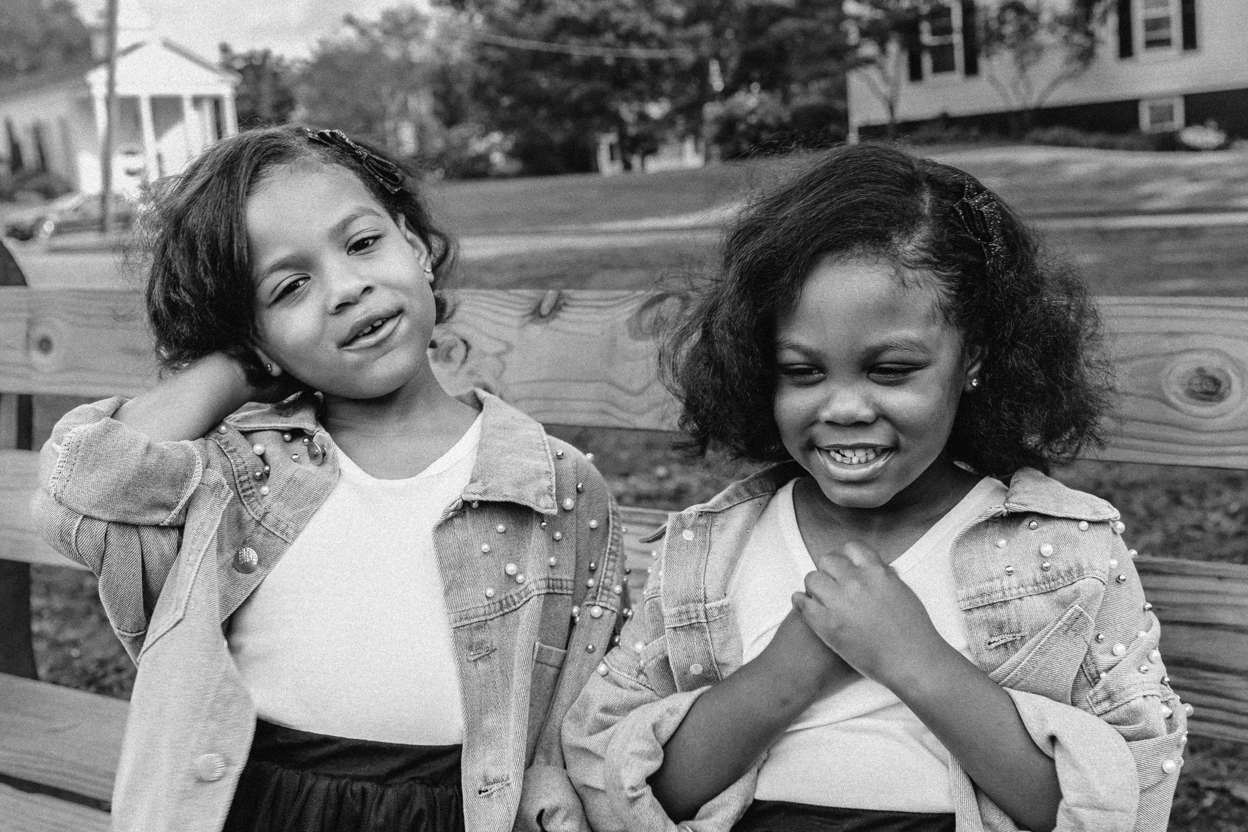 Two young girls with dark curly hair sitting on a park bench, wearing denim jackets with pearls, smiling and interacting in an outdoor setting. Documentary family photographer in New York City 