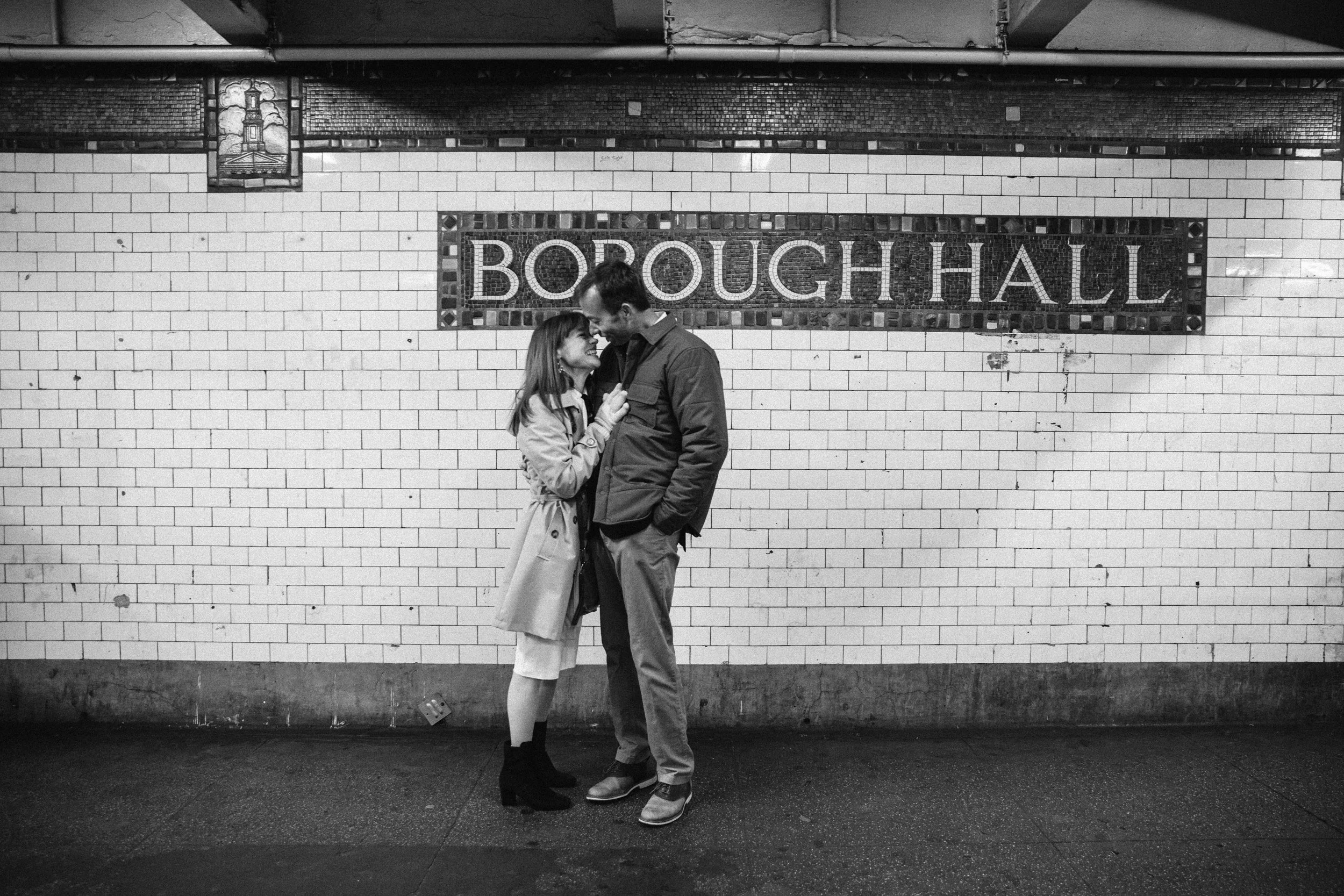A couple stands close together in a subway station with a sign reading 'BOROUGH HALL' in the background.