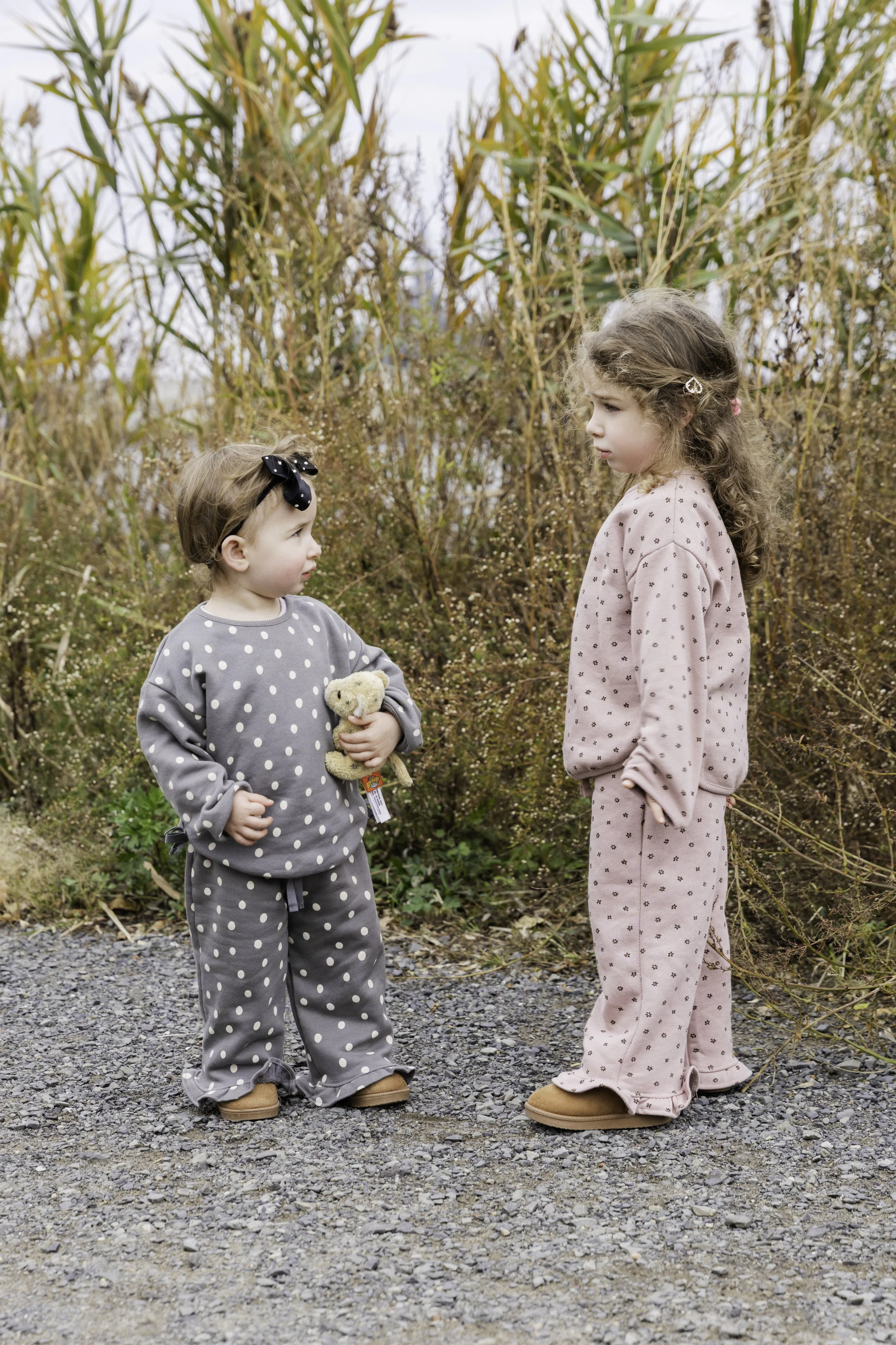 Two young girls standing outdoors on a gravel path, facing each other, with tall dry grass and bushes behind them.Outdoor family portrait session in NYC, candid and natural photography