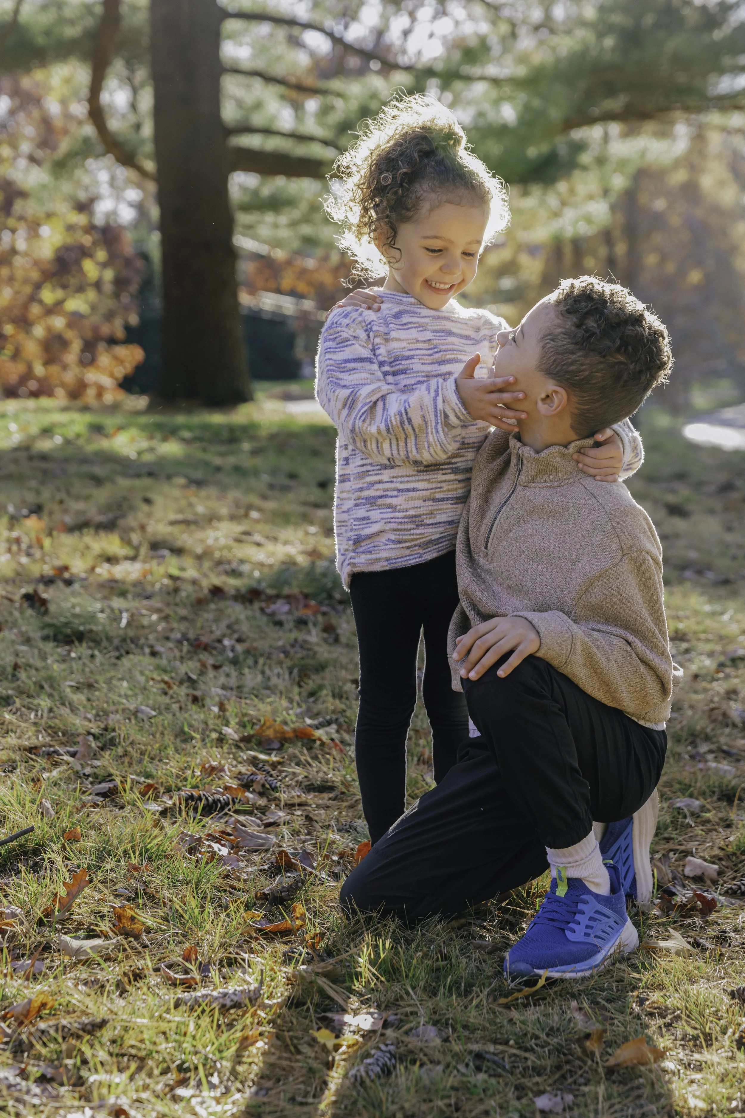 A young girl with curly hair smiling and touching the chin of a kneeling boy with short curly hair, Lifestyle family photography in New York City