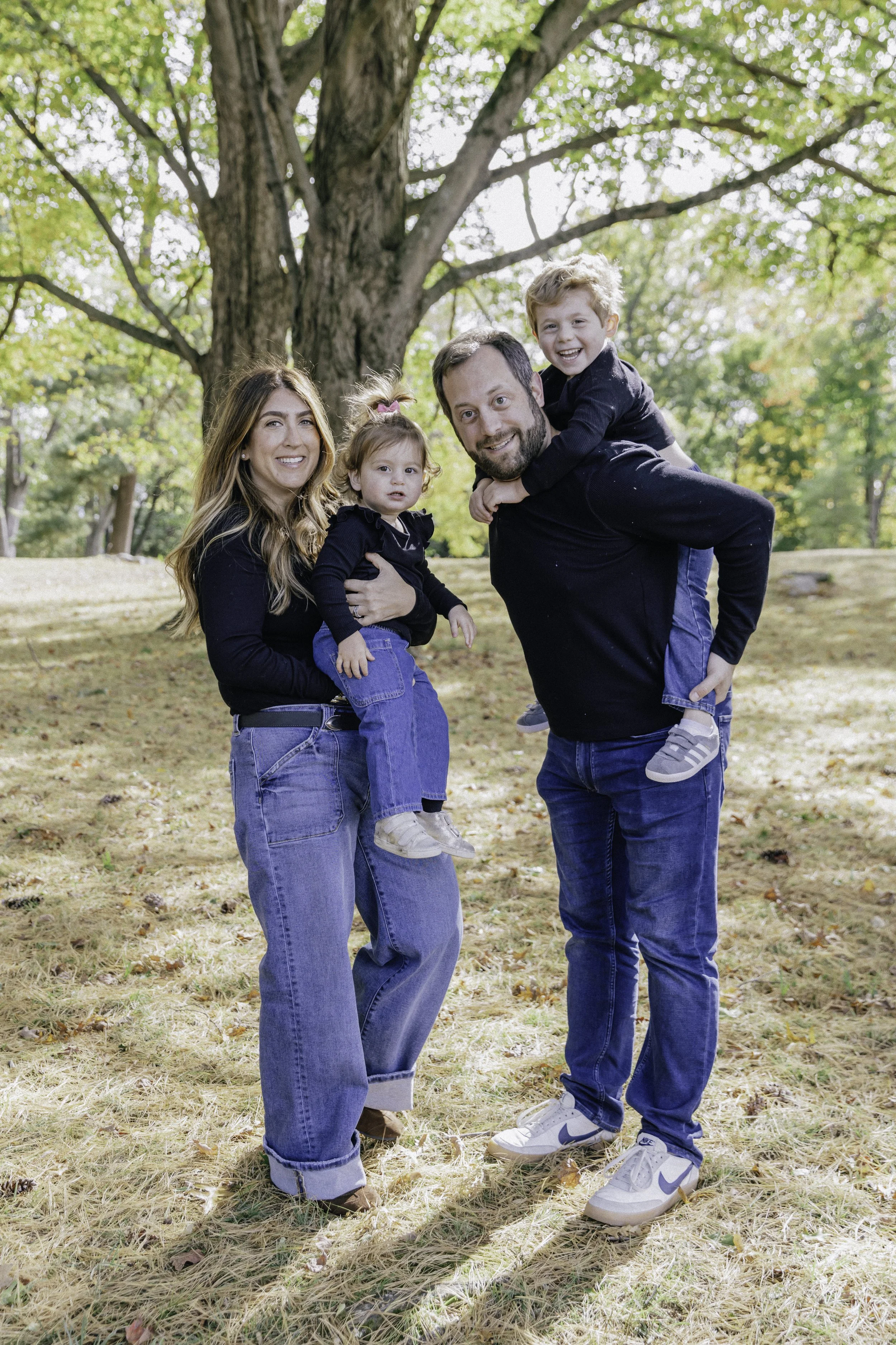 Family of four outside in a park with large trees, two adults and two children, smiling and playing.