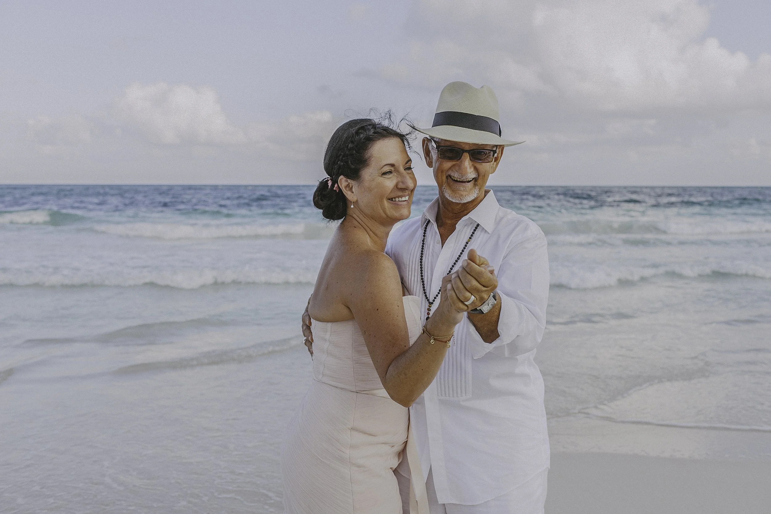 A happy couple dancing on the beach with the ocean in the background. The woman is wearing a light pink dress and the man is wearing white with a fedora hat and sunglasses.