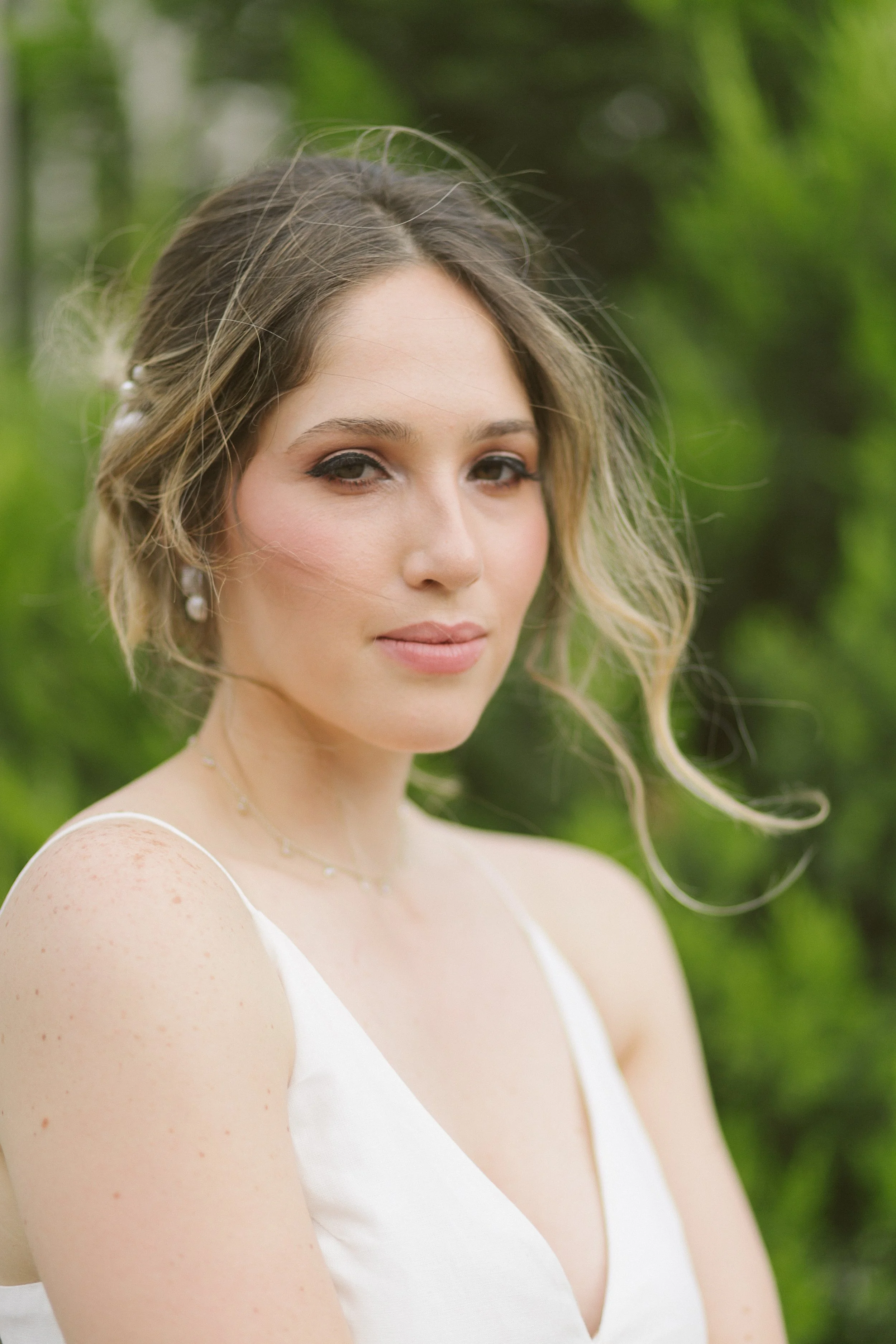 A woman with light skin, brown eyes, and wavy blonde hair wearing a white dress and jewelry outdoors with green foliage background.