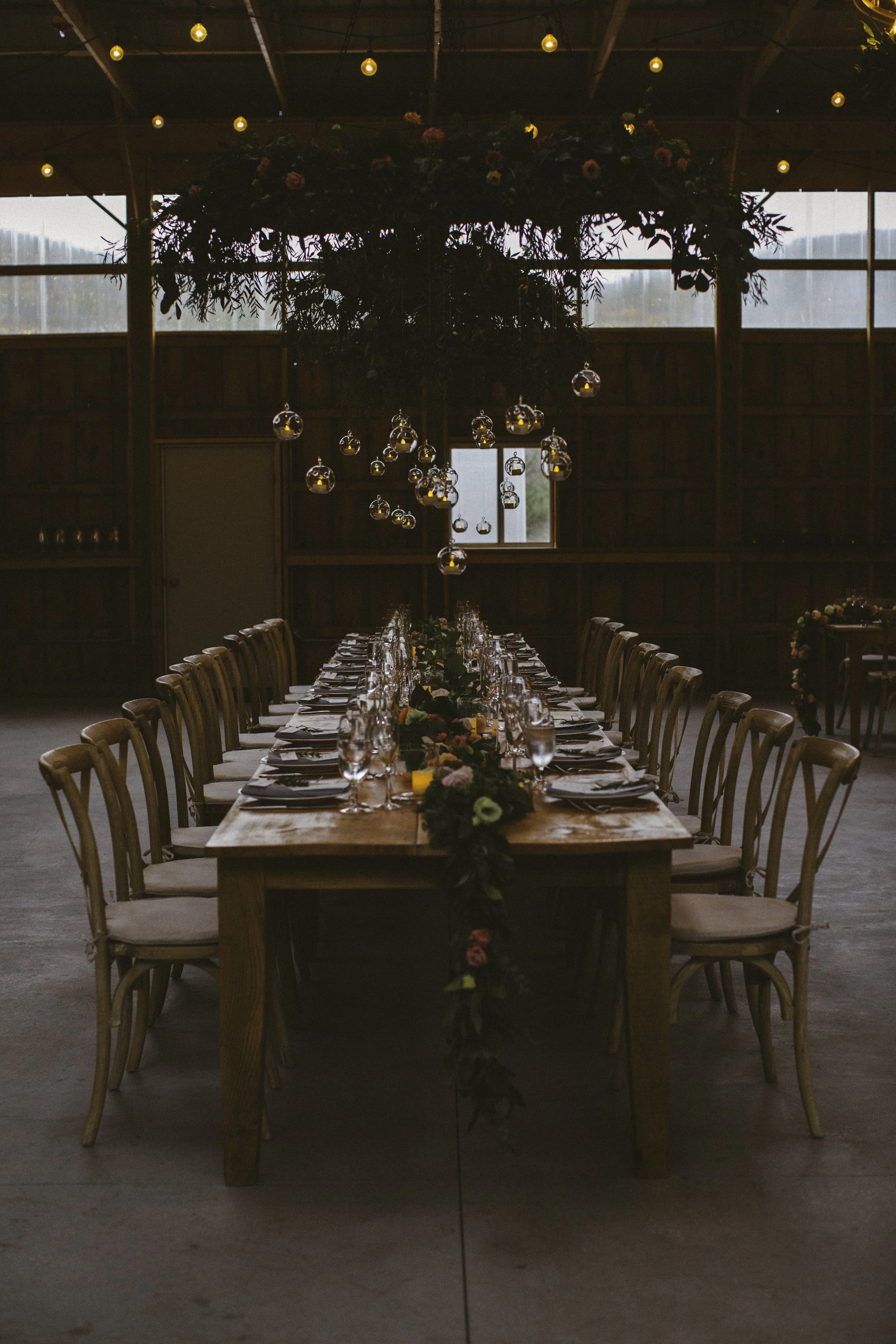 Long wooden banquet table set with glassware, cutlery, and floral table runner, decorated for a wedding or special event in a rustic barn with wooden walls and string lights hanging above, and a floral chandelier overhead.
