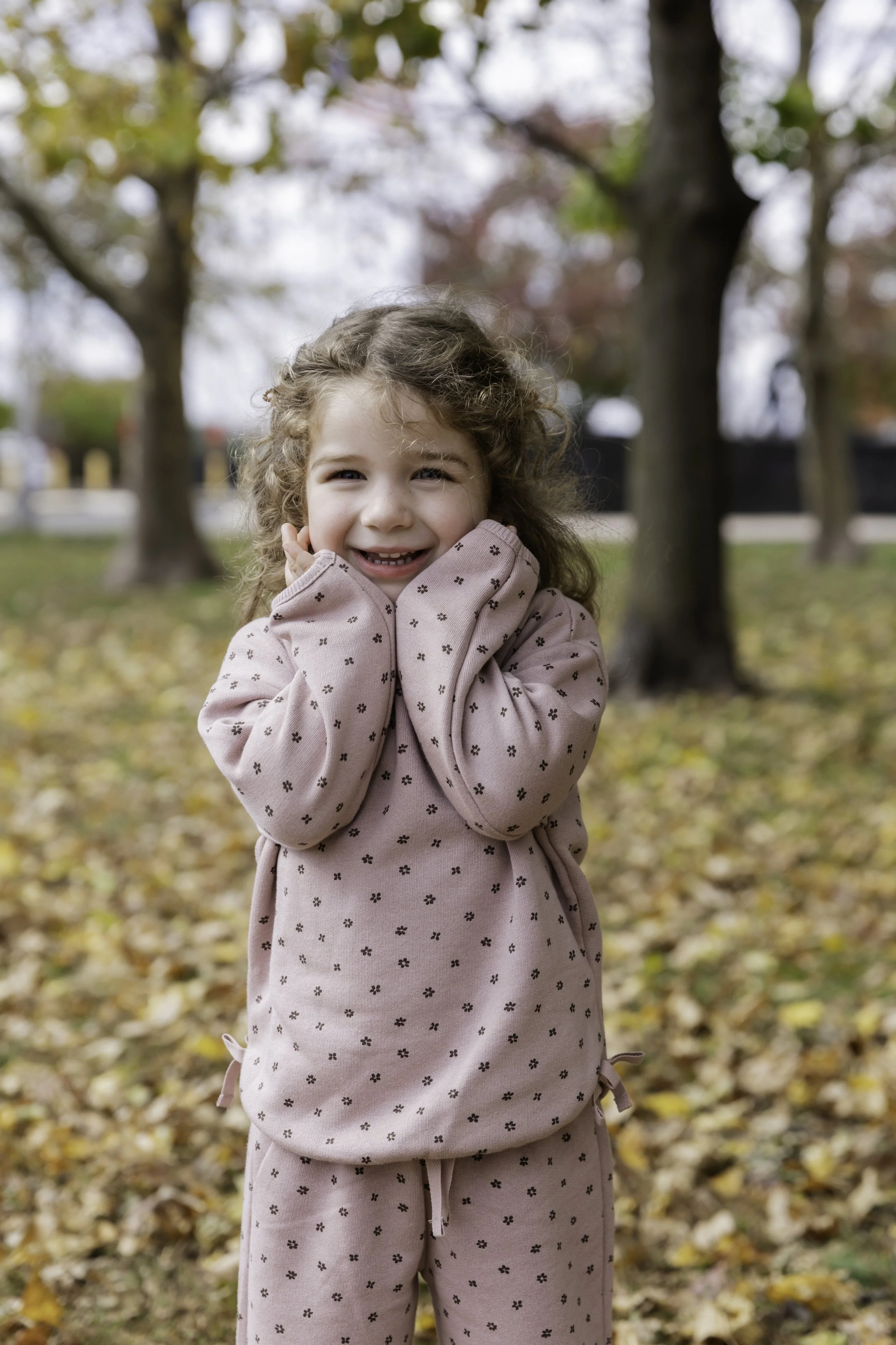 A smiling young girl with curly hair wearing a pink polka-dotted outfit standing outdoors among trees and fallen autumn leaves.