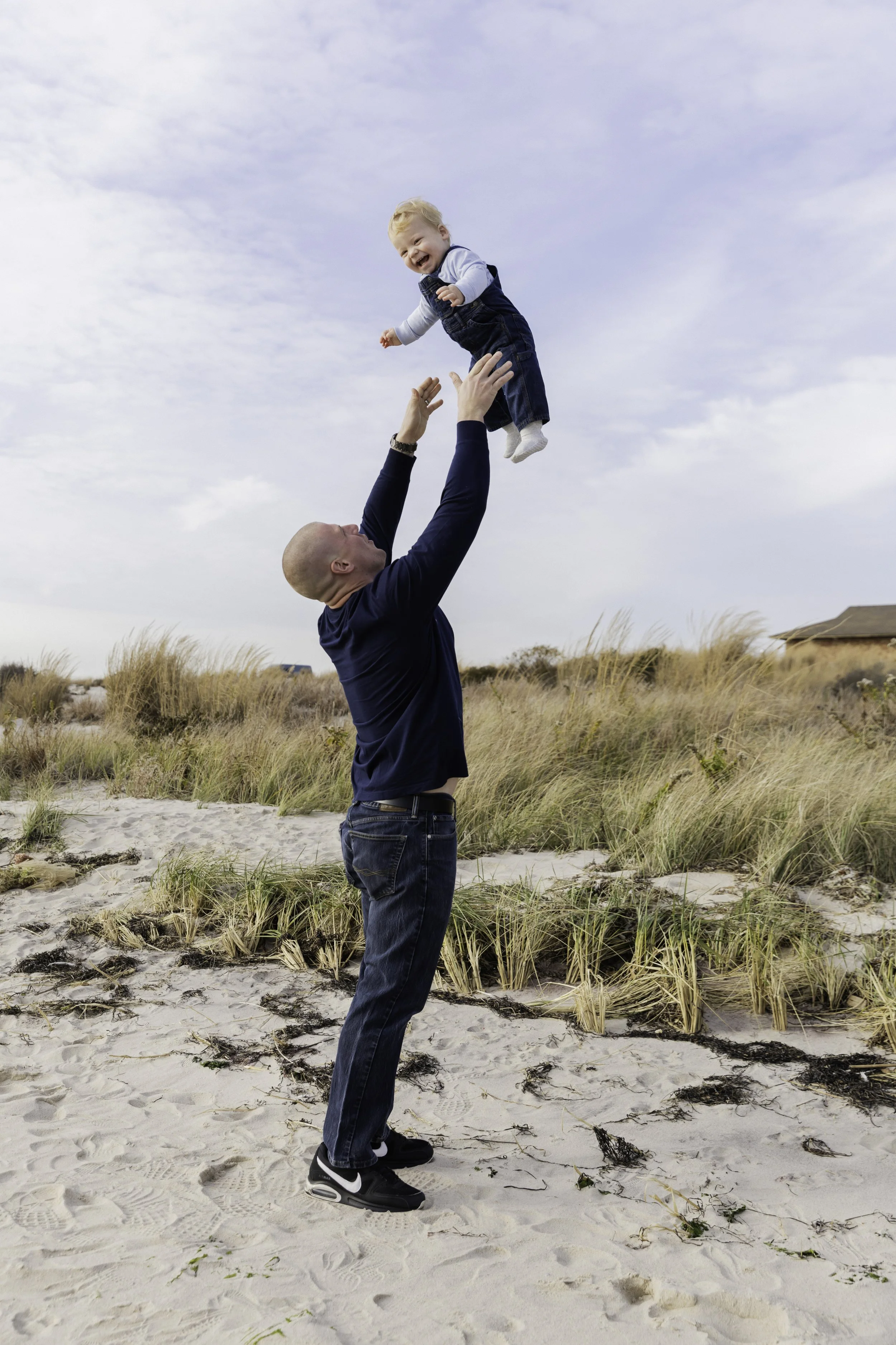 A man throwing a young child high in the air on a sandy beach with grass and a cloudy sky in the background. Documentary family photographer in New York City 