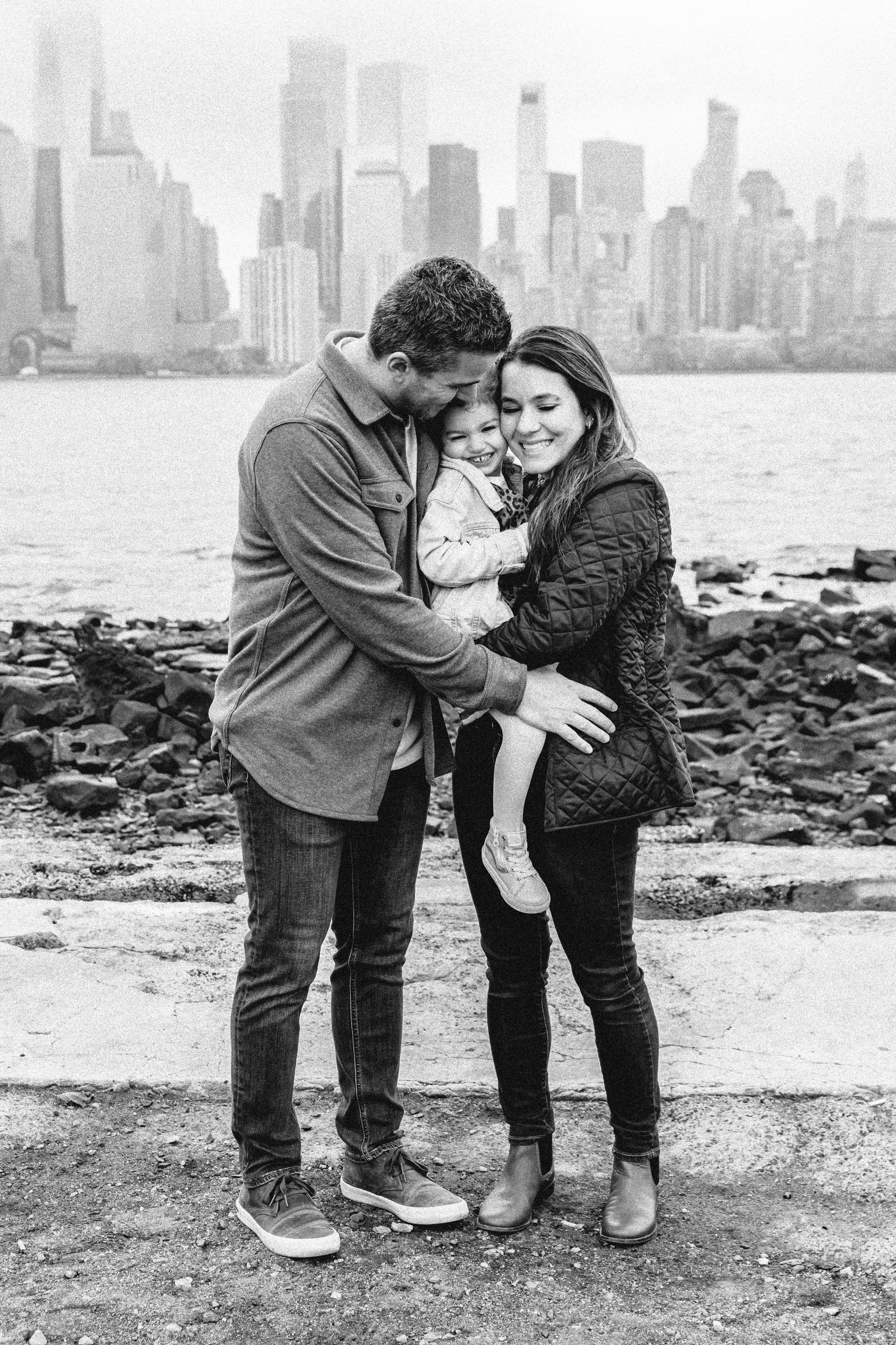 Family smiling by water with city skyline in background — NYC family photographer