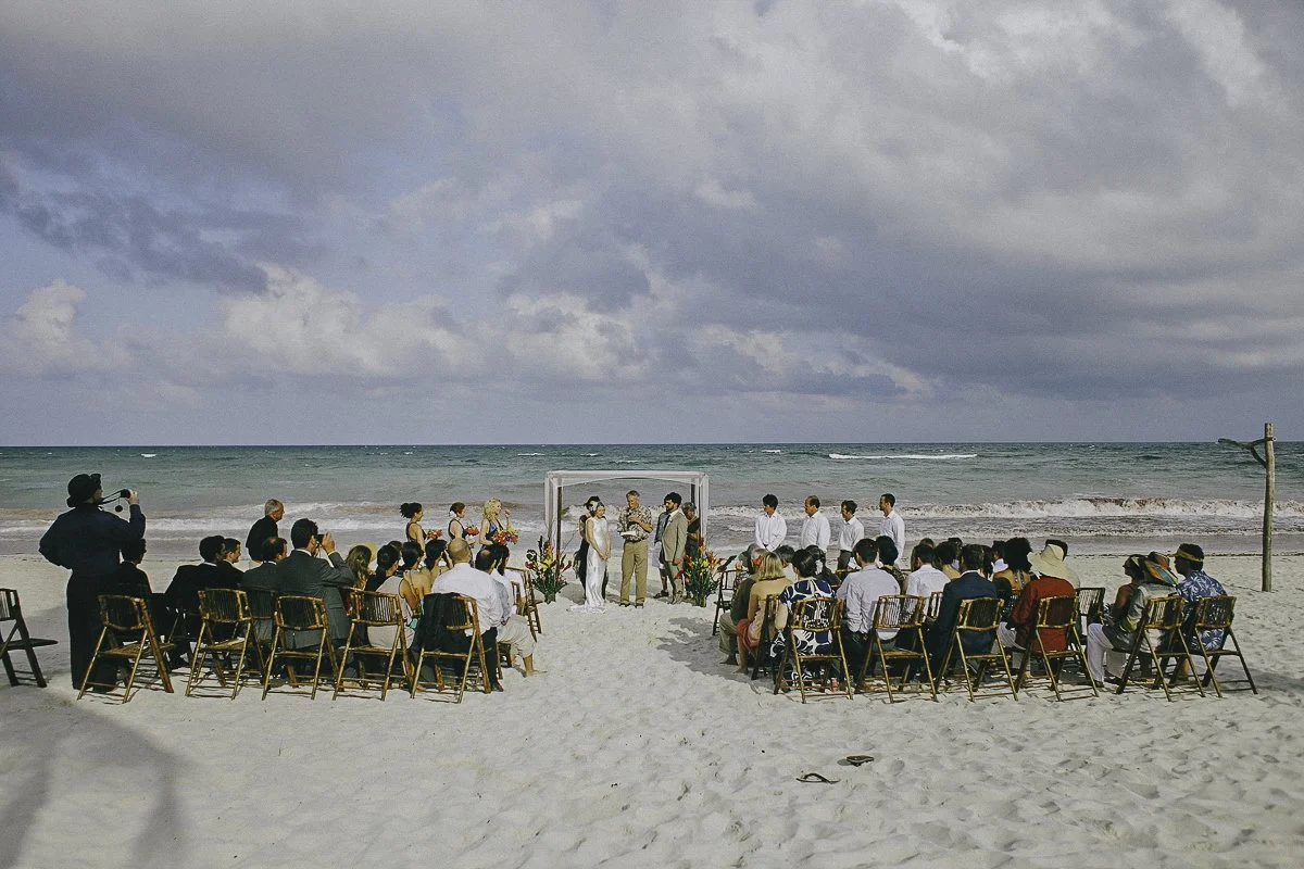 A beach wedding ceremony taking place on sandy shore with guests seated in chairs facing the ocean, and the sea and cloudy sky in the background.