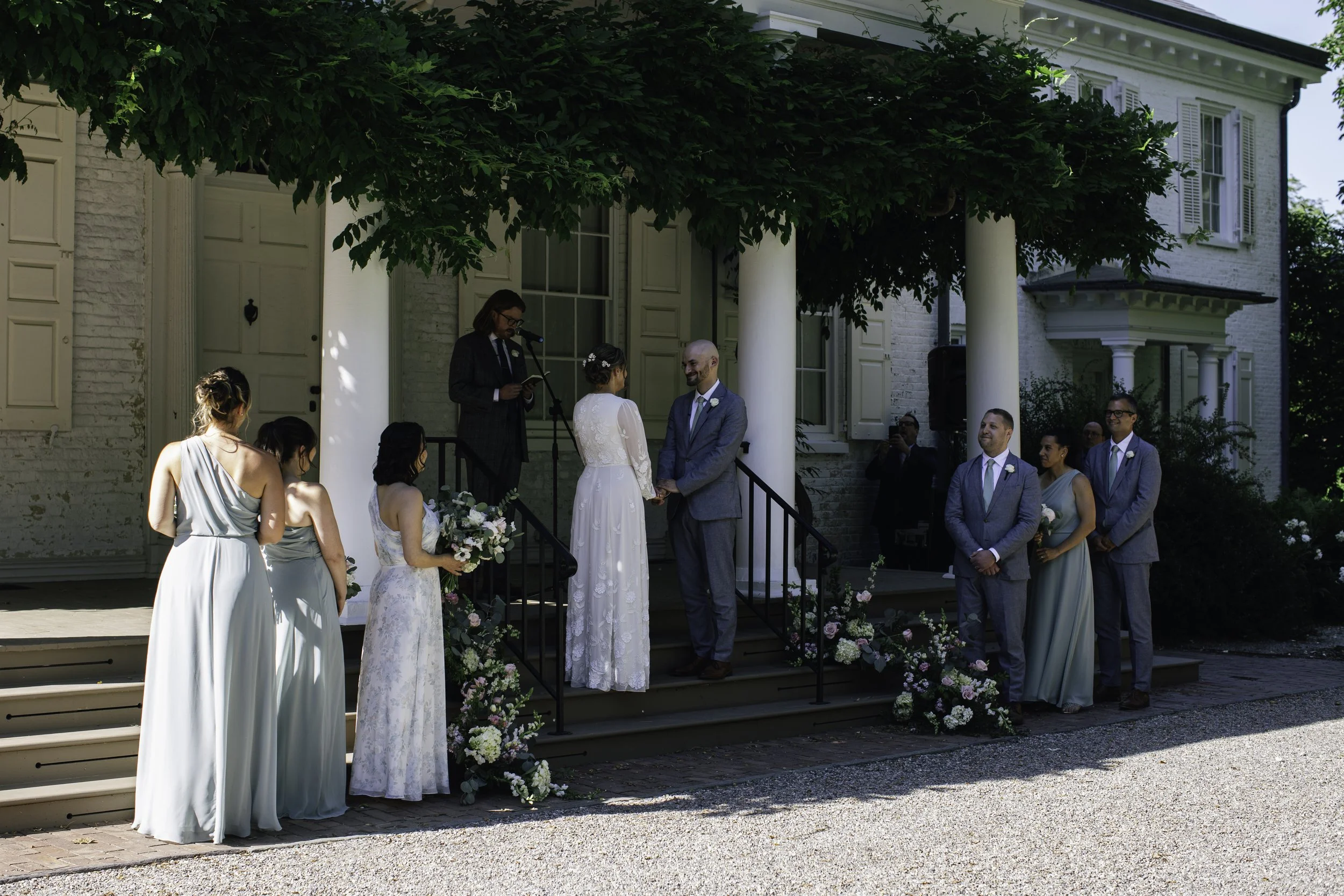 A wedding ceremony taking place outdoors in front of a white house with greenery. The bride and groom stand on a small porch, holding hands, with a woman officiating the ceremony reading from a book. Bridesmaids and groomsmen stand nearby, and floral