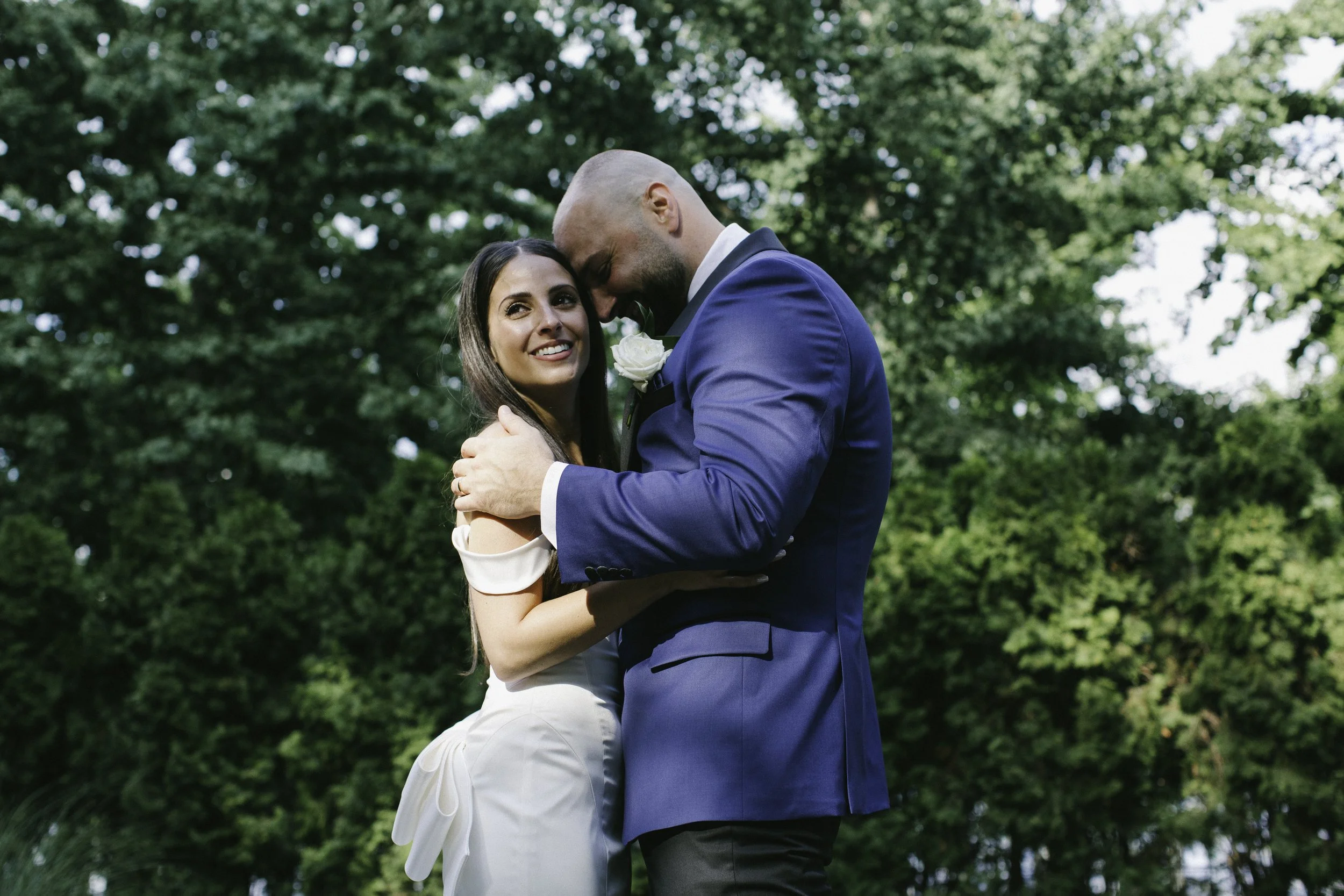 A newlywed couple sharing an intimate moment outdoors, with the man in a blue suit and the woman in a white wedding dress, surrounded by green trees. Documentary wedding photography in NYC