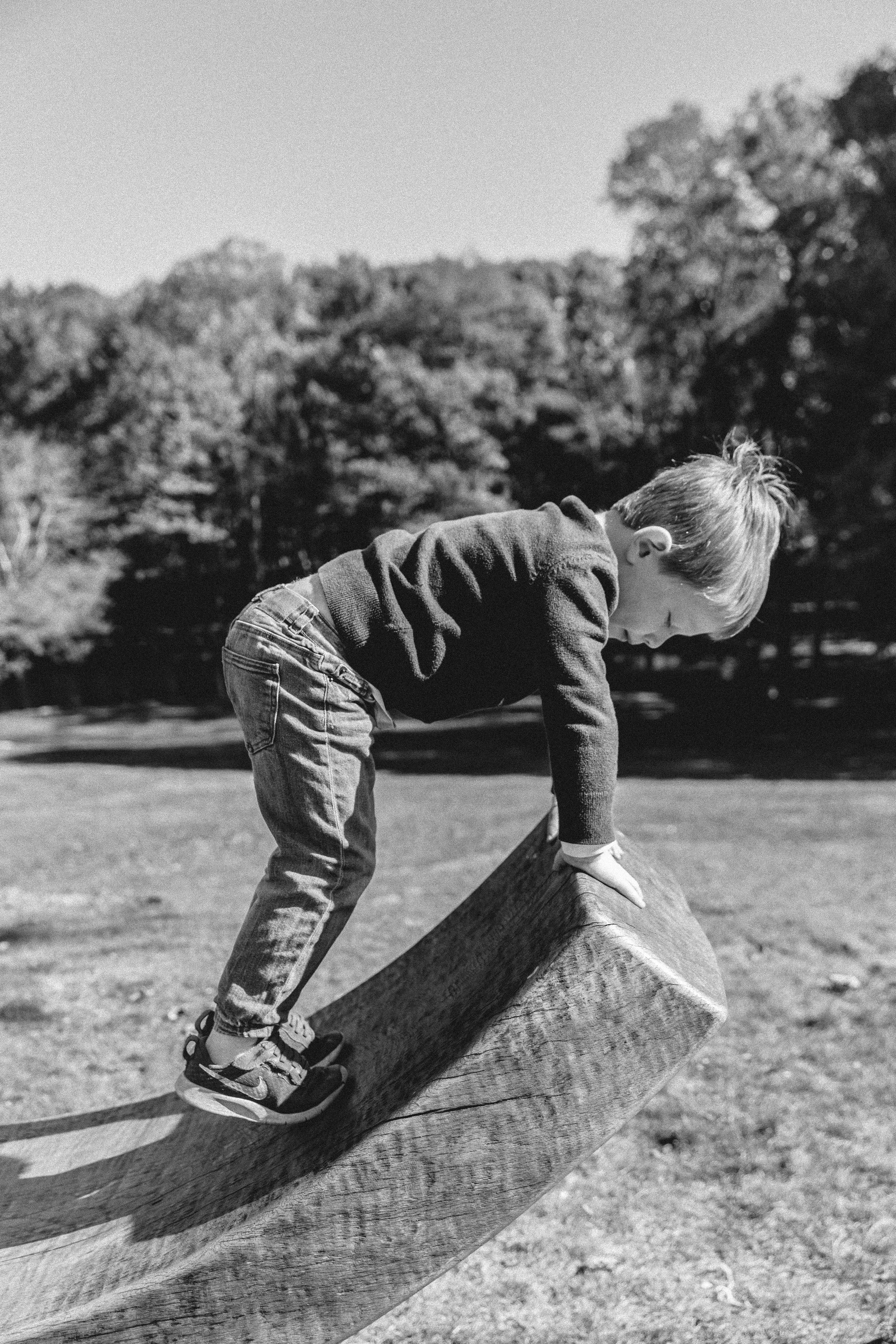 A young boy climbing on a large wooden log in a park with trees in the background. Outdoor family portrait session in NYC, candid and natural photography