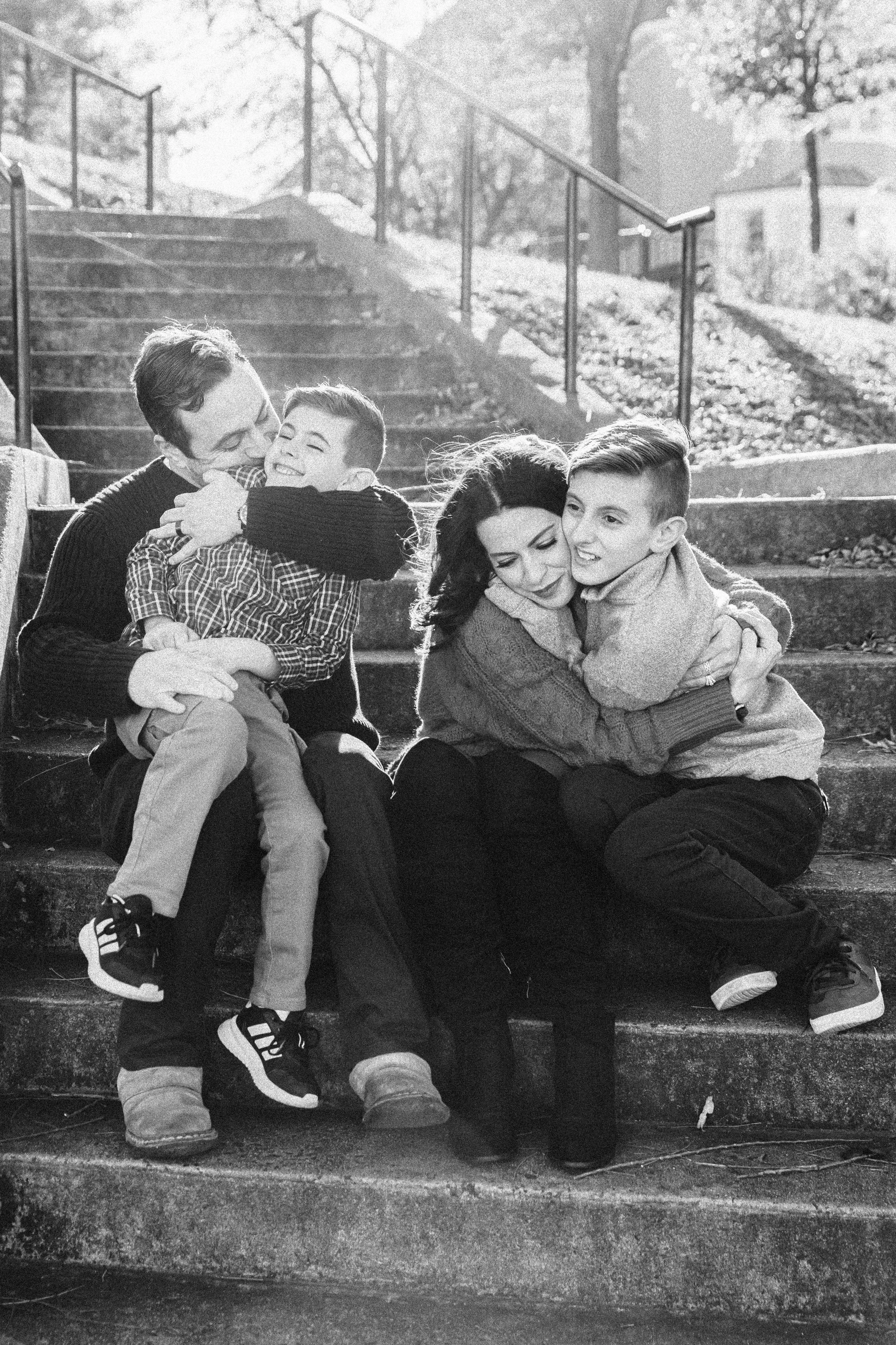 A black and white photo of a family sitting on outdoor stairs, hugging and smiling. Lifestyle family photography in New York City