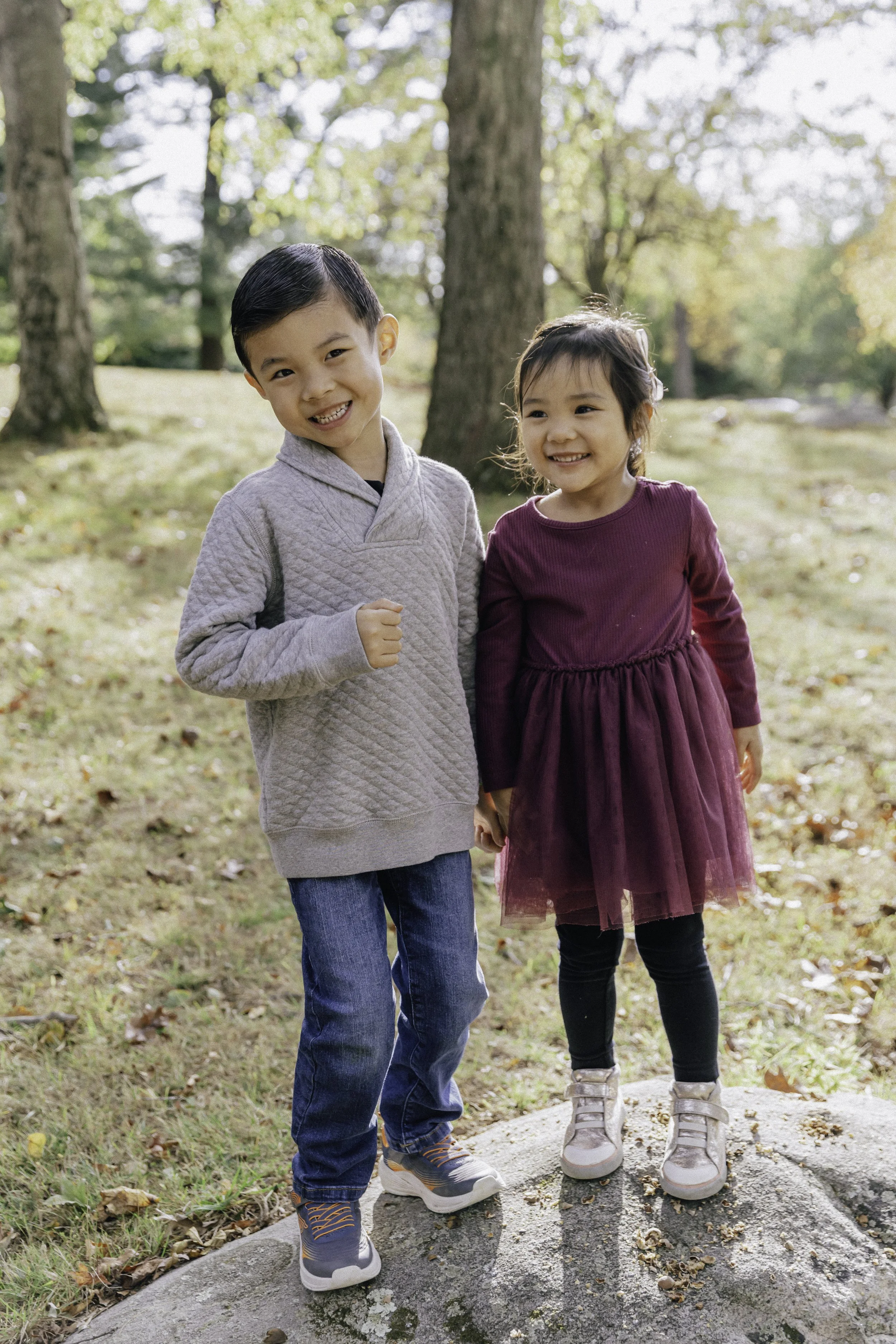 Two kids, a boy and a girl, holding hands and smiling outdoors in a park with fall foliage.
