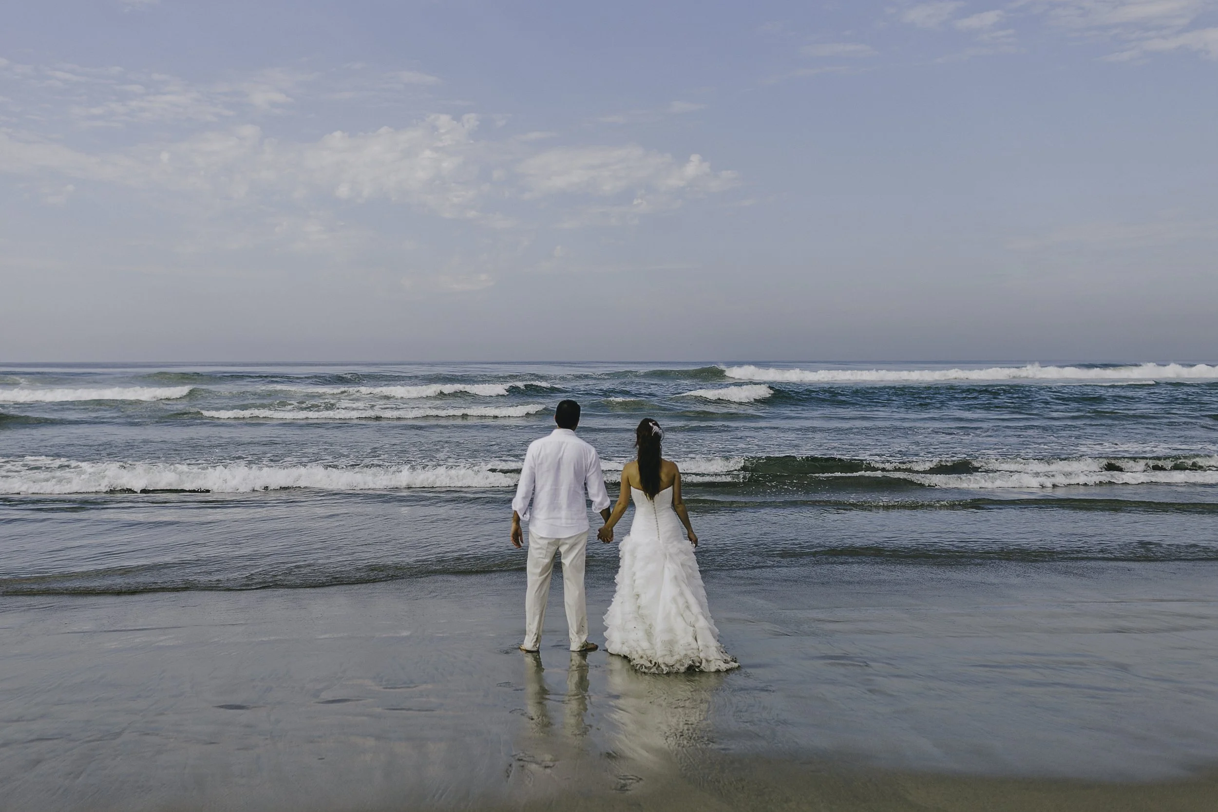 A couple in wedding attire holding hands standing on the beach, facing the ocean under a partly cloudy sky. NYC wedding photography
