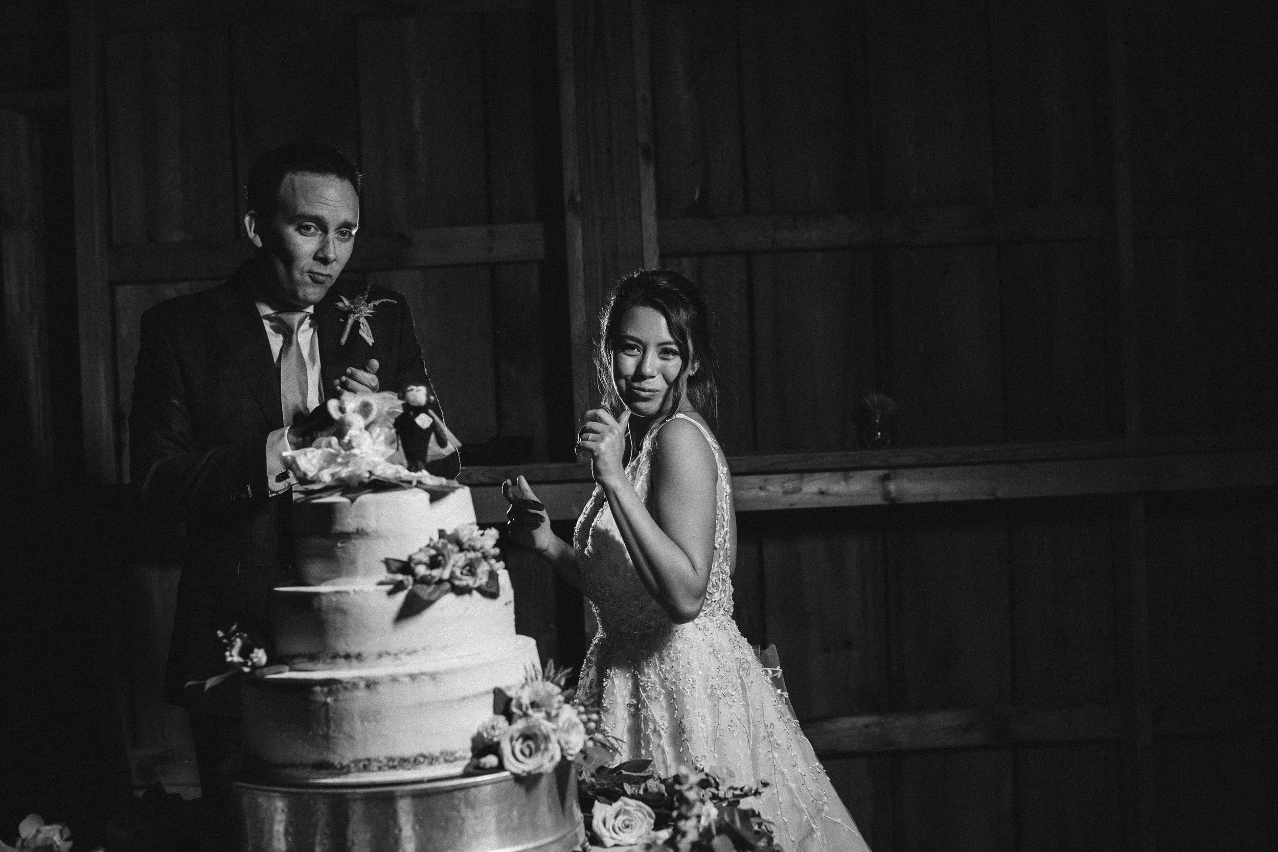 Black and white photo of a bride and groom at their wedding. The groom is standing near a large, tiered wedding cake with floral decorations and bride and groom figurines on top. Documentary wedding photography in NYC