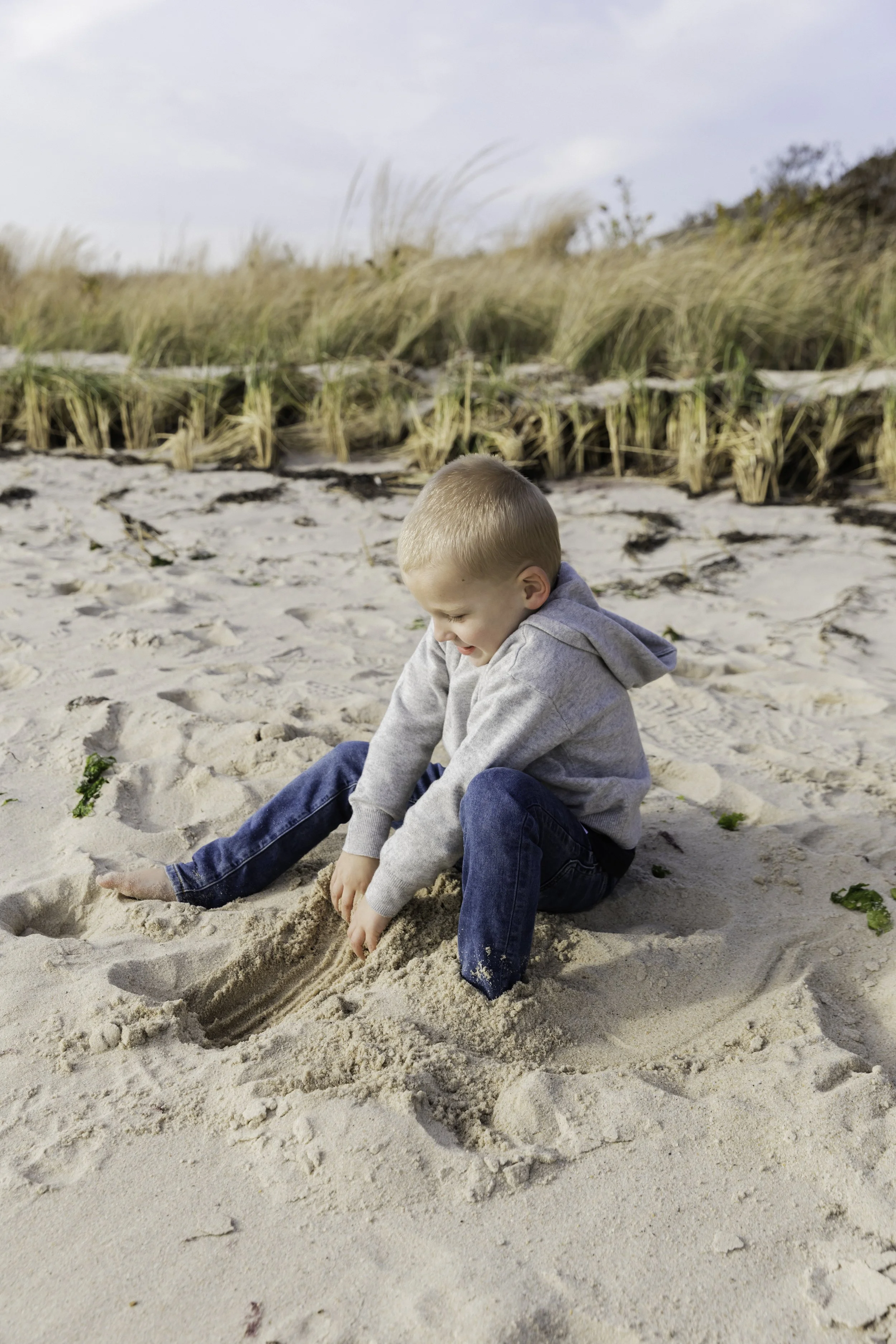 A young boy playing in the sand on a beach, wearing a grey hoodie and jeans, with tall grasses and a cloudy sky in the background.