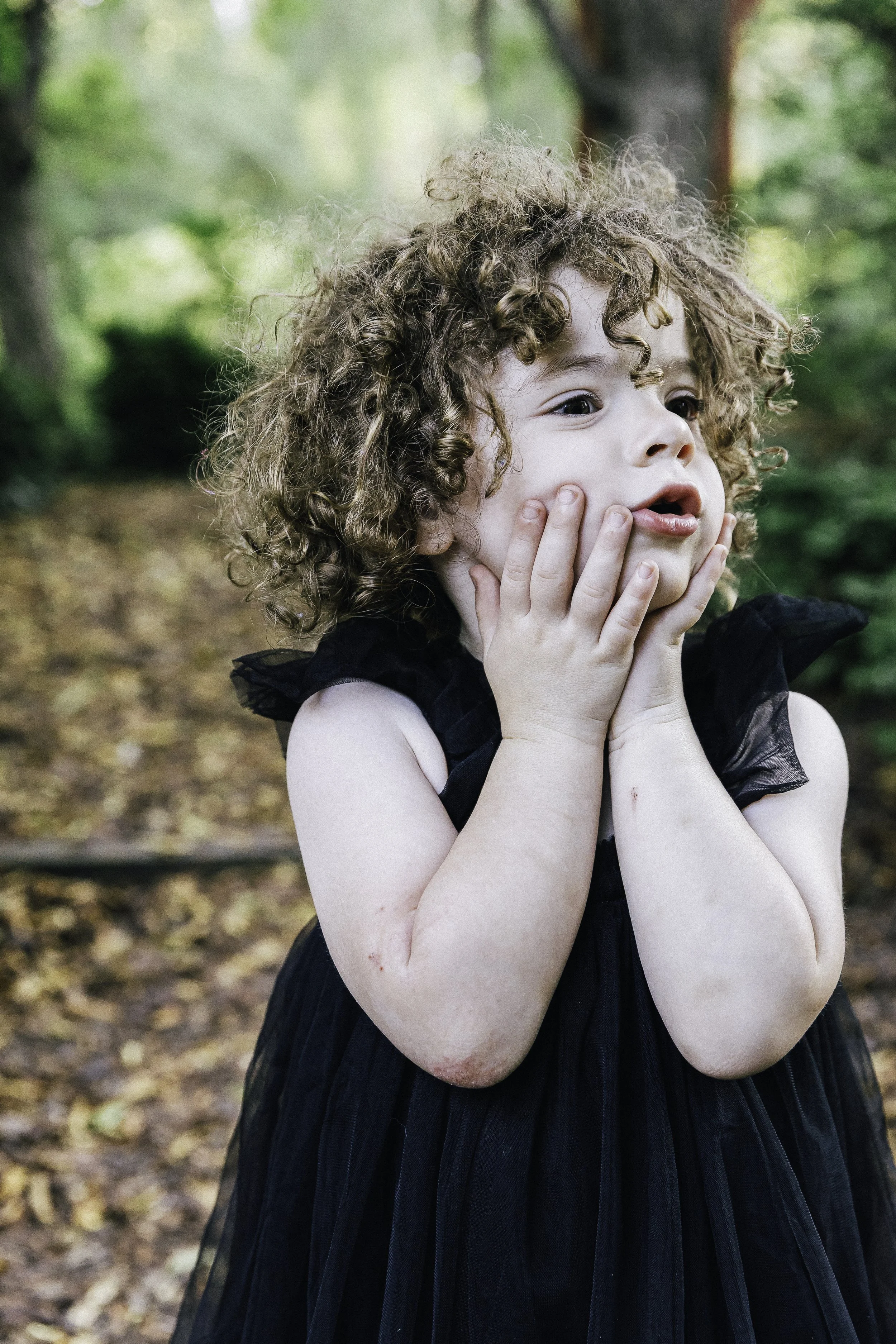 A young girl with curly hair, wearing a black dress, holding her face with both hands, standing outdoors in a wooded area.