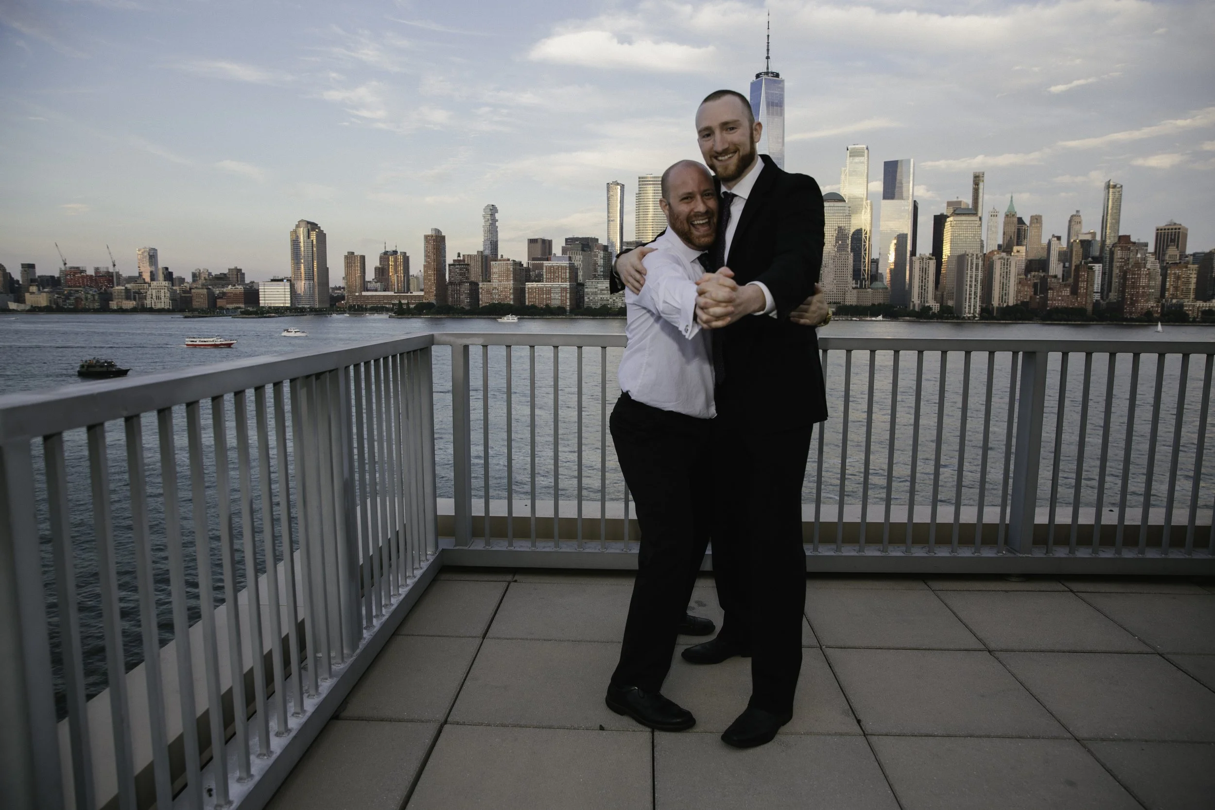Two men in formal attire hugging on a balcony with a city skyline and water in the background.